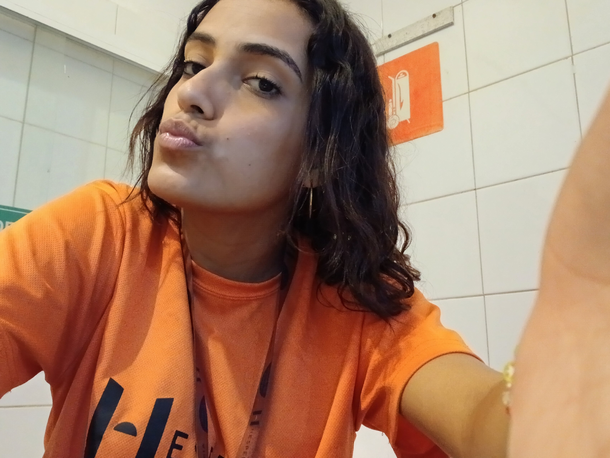 A young woman with warm skin tone and dark, curly hair is featured in a close-up selfie, captured from a slightly low, upward angle. She is looking directly at the camera with a playful expression, puckering her lips in a "duck face." A small mole is visible on her left cheek (viewer's right). She wears a bright orange, textured t-shirt with dark, partially visible text on the front; parts of letters like "H" and "E" are discernable. A dark lanyard or strap is visible around her neck, with faint, illegible text. She has a slender, gold-colored hoop earring in her right ear (viewer's left) and a light-colored, possibly beaded, bracelet on her right wrist, which is partially visible on the right side of the frame as she holds the camera.
The background consists of light-colored, possibly white, tiled walls, suggesting an indoor or semi-enclosed environment. On the wall behind her, to the right, there is a rectangular orange sign with a white graphic of what appears to be a two-wheeled cart or bin. Above this sign, a horizontal metal bar or fixture is attached to the wall. Another green sign is partially visible on the far left, though its details are obscured. The overall lighting suggests an indoor setting, and the clean tiled walls hint at a public or service area within Playa El Agua, Venezuela.