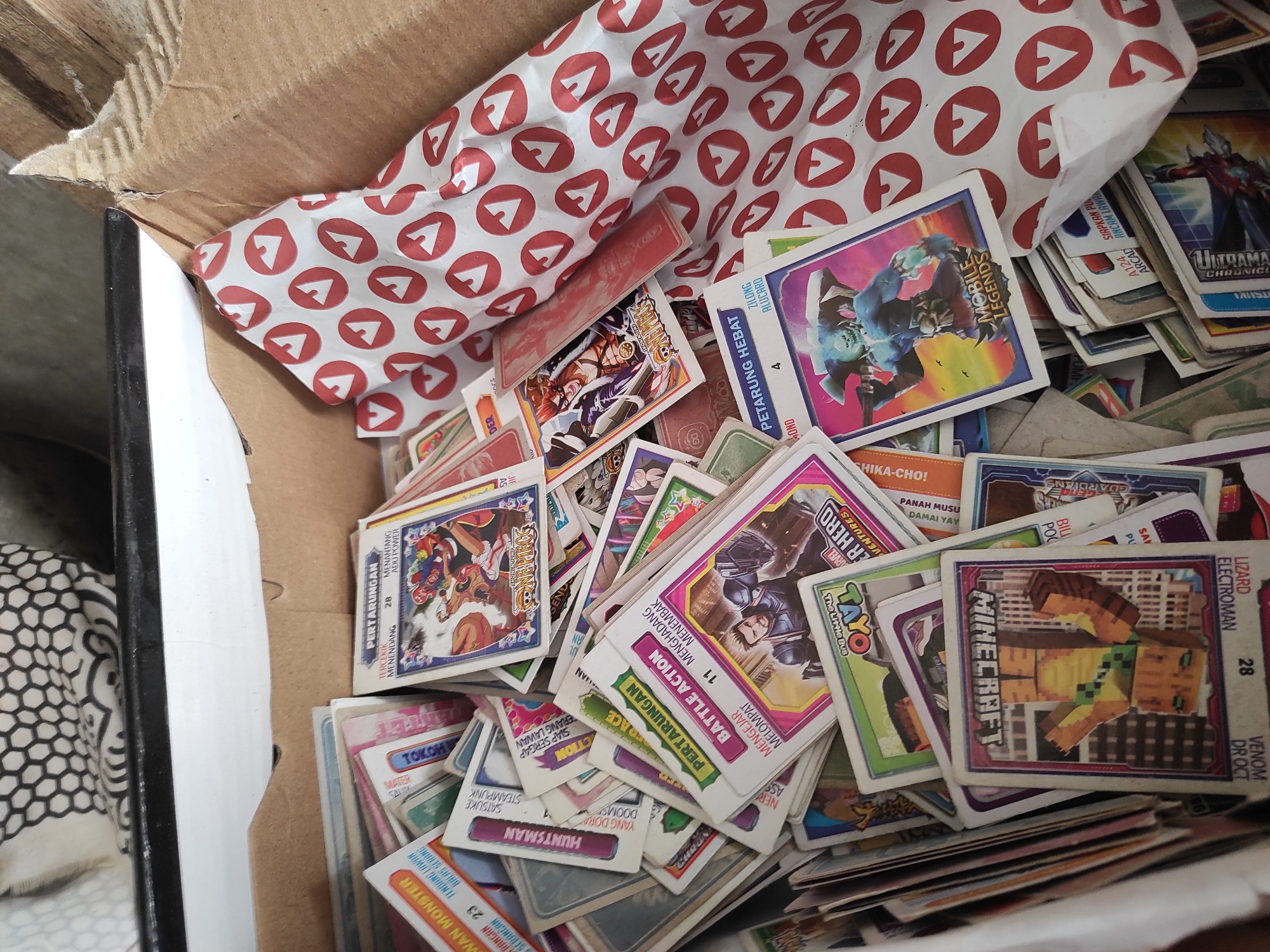 In the city of Sumedang, Indonesia, a cardboard box is filled with a collection of trading cards. The cards feature various characters and designs, including images from "One Piece," "Mobile Legends," "Minecraft," and what appear to be generic martial arts or monster-themed fighters. Some cards have text in Indonesian, such as "PERTARUNGAN" (Fight), "PETARUNG HEBAT" (Great Fighter), and "BATTLE ACTION." There is also a piece of patterned paper with red circular symbols, likely wrapping paper, also inside the box. The overall impression is of a well-loved collection of game and anime-related trading cards.