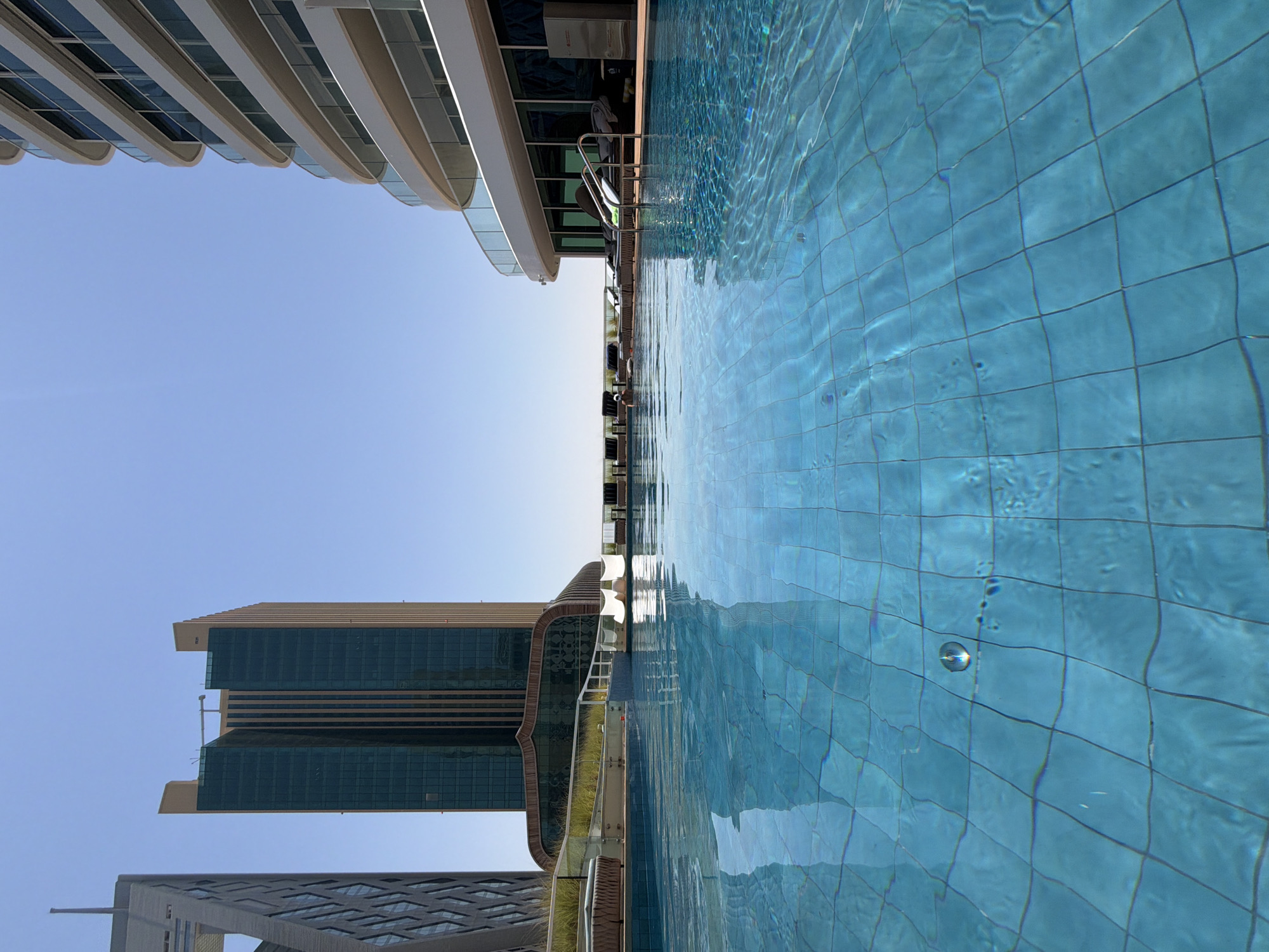 An outdoor swimming pool with turquoise water, tiled in a grid pattern, is in the foreground. Sunlight glints off the water's surface, creating ripples and reflections. In the background, several modern skyscrapers dominate the skyline under a clear blue sky. To the left, a tall, angular building with a patterned facade is visible. In the center, a rectangular skyscraper with a dark green glass facade rises high. To the right, a curved, multi-story building with a white exterior and balconies featuring glass railings is prominent. Around the pool, lounge chairs are arranged on a wooden deck, and some are occupied by people relaxing. A white water slide is positioned at the far end of the pool area.