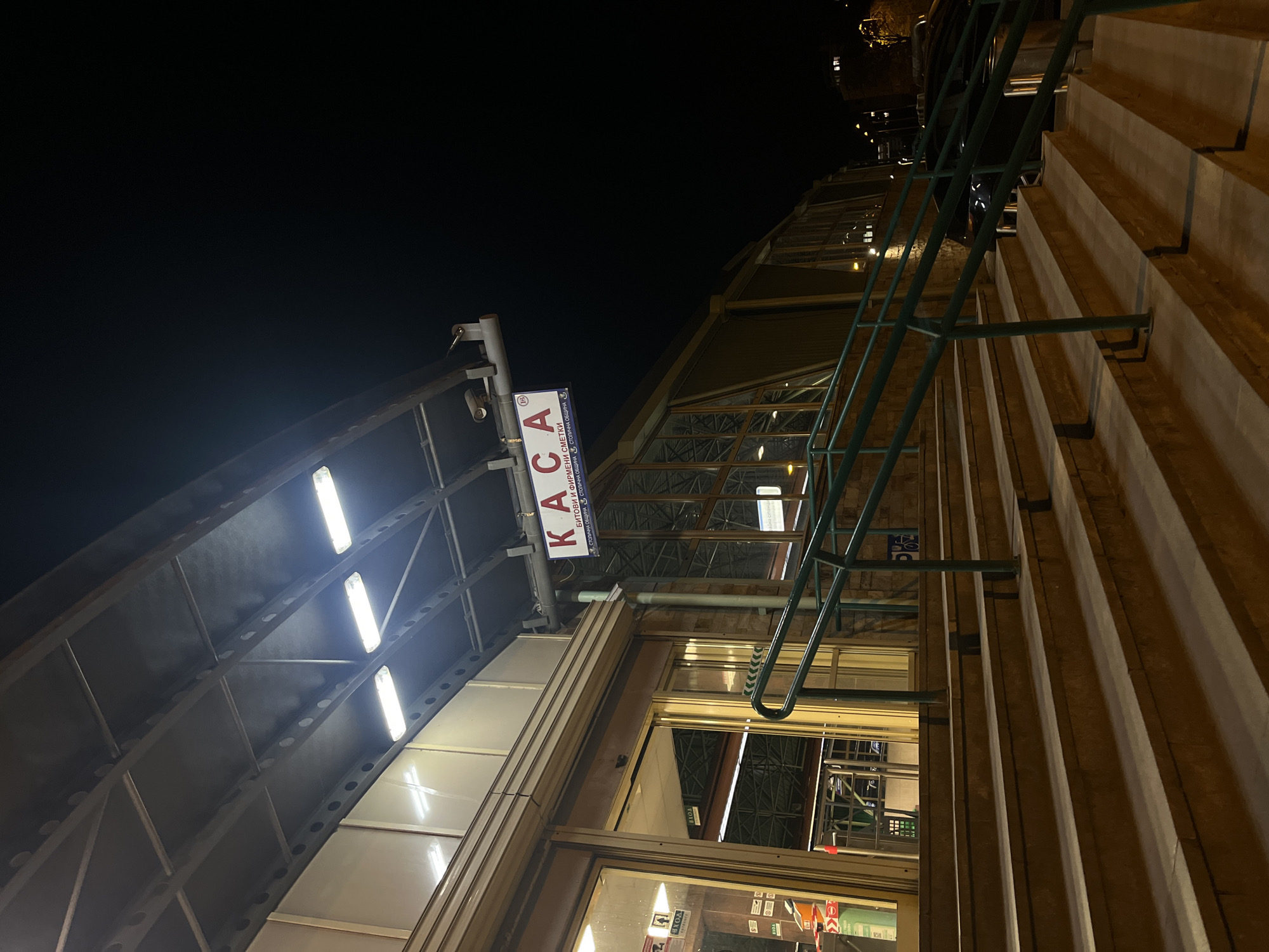 The image captures an upward-looking night view of an urban architectural structure, likely in Sofia, Bulgaria. The sky is dark, indicating nighttime, with strong artificial lighting illuminating the scene.
On the left, a covered walkway or entrance features a series of bright, elongated fluorescent lights embedded in its ceiling structure. Suspended from this canopy is a rectangular white sign with prominent red Cyrillic text reading "KACA" (KASA), meaning "Cashier" or "Box Office." Below this, smaller Cyrillic text is visible, stating "БИТОВИ И ФИРМЕНИ СМЕТКИ" (Household and Business Bills) and "СТОЛИЧНА ОБЩИНА" (Sofia Municipality).
To the right, a multi-story building rises with a textured facade, appearing to be made of concrete or stone with horizontal ridges. A distinct green metal railing or ladder-like structure runs vertically along this building. The building features large glass windows, through which interior lighting and some structural elements, possibly a metal trolley or scaffolding, are visible. A small blue sign with a wheelchair accessibility symbol is also discernible through the glass.
The perspective of the photograph is angled upwards, giving a sense of the height and scale of the structures. No people are present, and no specific activity is taking place beyond the ambient operation of the building and its lighting. The overall impression is that of a municipal service center or payment office, open during the evening hours.