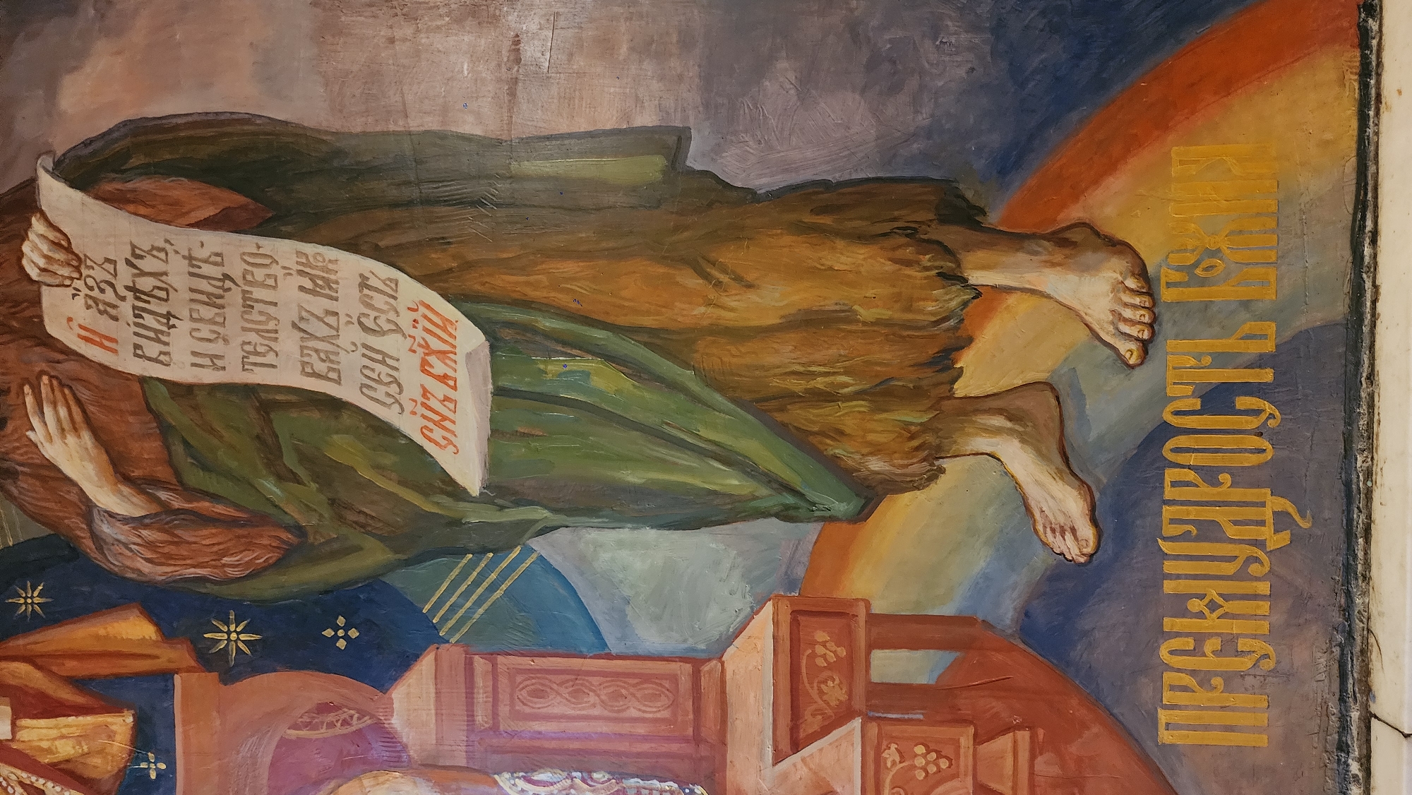 The image shows a section of an Eastern Orthodox religious painting. A figure, draped in brown and green cloth, floats horizontally. Their hands hold a scroll inscribed with text in Cyrillic letters, some of which are highlighted in red. The figure's feet point right. To the right of the figure, vertically stacked Cyrillic letters in gold spell out words. The background features a rainbow arc and geometric shapes. Below, there is an image of wooden furniture. The painting shows signs of age and wear.