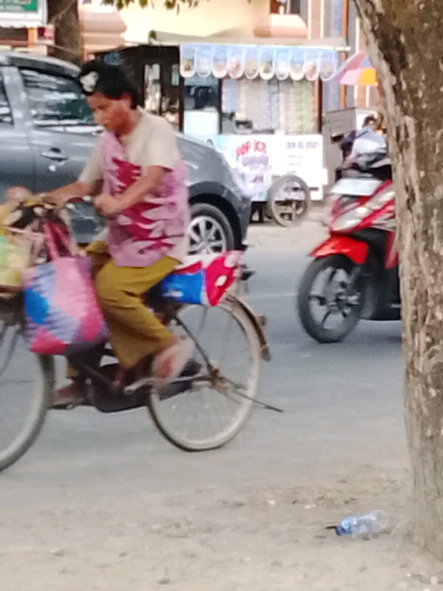 The image depicts a street scene in Slawi, Indonesia. A person is riding a bicycle from left to right across the frame. The cyclist is wearing a light-colored t-shirt with a pink and red pattern and yellow pants. They are carrying a colorful bag on the front of the bicycle. In the background, there is a red and black motorcycle parked, and further back, a food cart with an awning. There is also a silver car visible on the left side of the image. A tree trunk is prominent on the right side of the image, partially obscuring the view. The ground is a mixture of pavement and dirt.