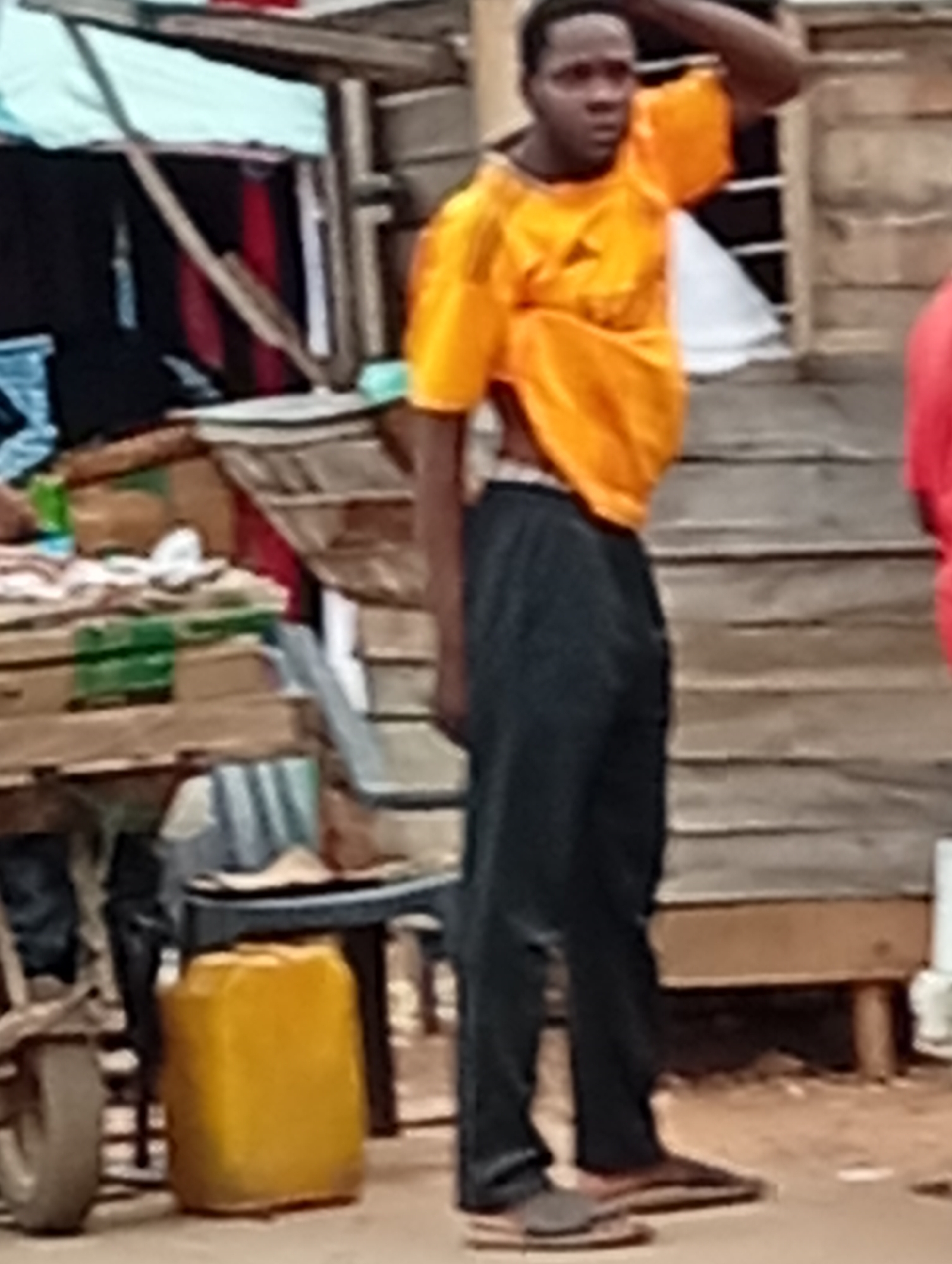 A person is standing next to a wooden structure that appears to be a market stall in an outdoor urban environment. The person is wearing an orange t-shirt and black pants. In the foreground, there is a yellow container. Behind the person, there are shelves with various items, including boxes and what looks like clothing. The ground is dirt. The lighting suggests it is daytime.