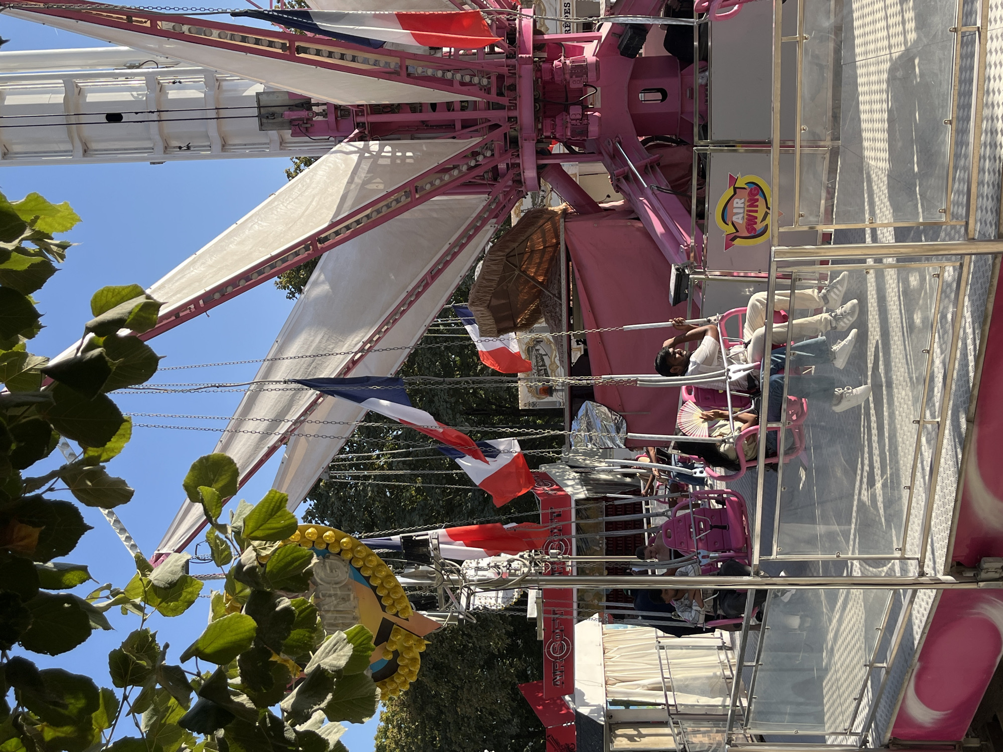 The image captures a lively scene at a funfair or amusement park, likely located in Paris, France, given the presence of French flags adorning a ride. The focal point is a large, pink amusement ride, possibly a swing ride, with white fabric sails extending outwards. The ride has numerous chains and appears to be in operation, with several people seated on swinging seats, enjoying the experience. French flags are prominently displayed, adding a patriotic touch to the festive atmosphere. In the foreground, trees with green leaves partially obscure the view, suggesting an outdoor setting with natural elements integrated into the park's landscape. The ground beneath the ride is a checkered metal platform, and the overall impression is one of gaiety and excitement, characteristic of a day at an amusement park. The scene is illuminated by bright sunlight, casting shadows and highlighting the vibrant colors of the ride and flags.