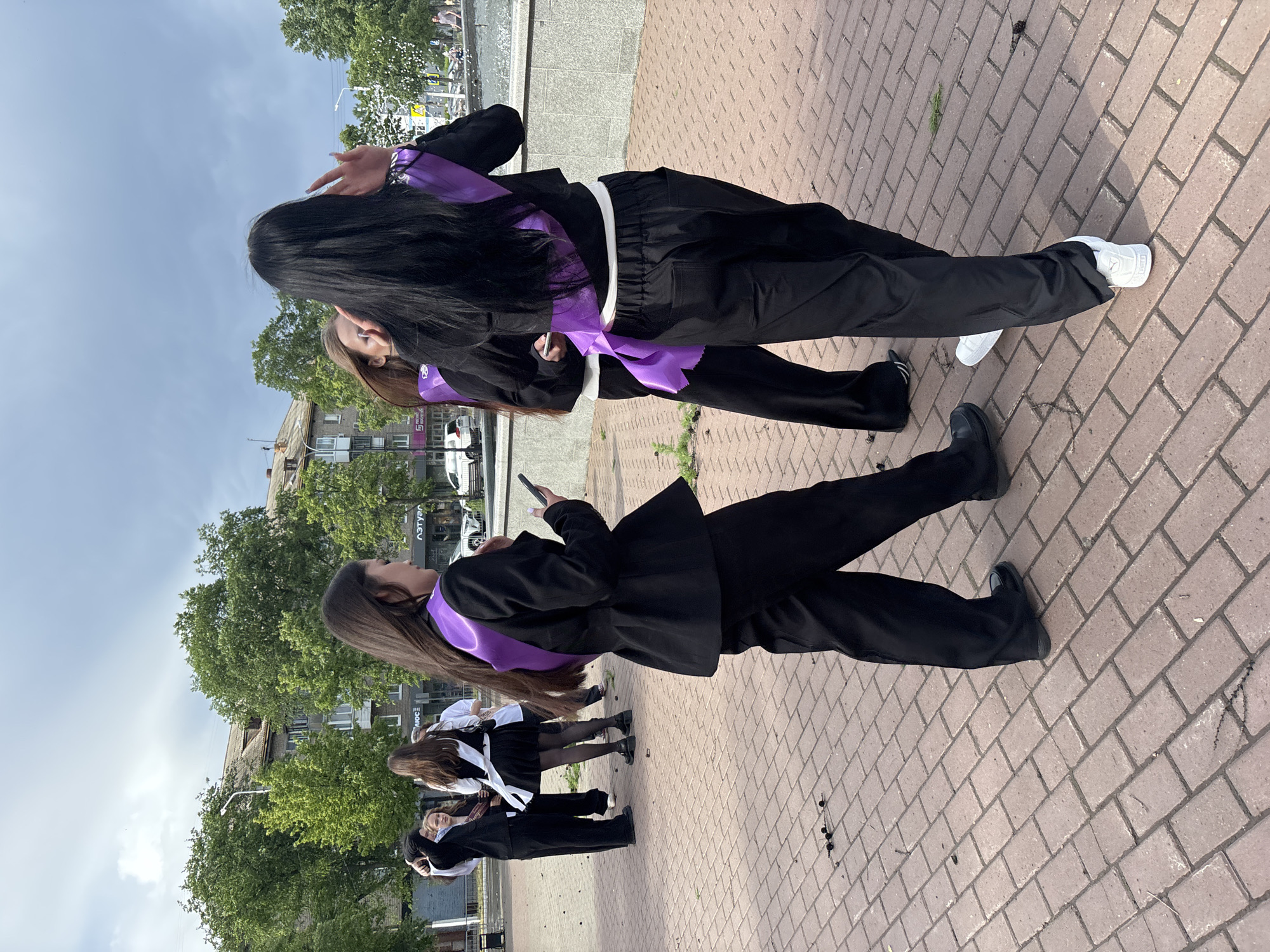 The image shows a group of people, presumably graduates, standing on a brick-paved surface. They are wearing black suits with purple and white sashes. The scene appears to be in an urban environment, with buildings and trees visible in the background. One person is looking at a phone. The location is Chaikovsky, Russia.