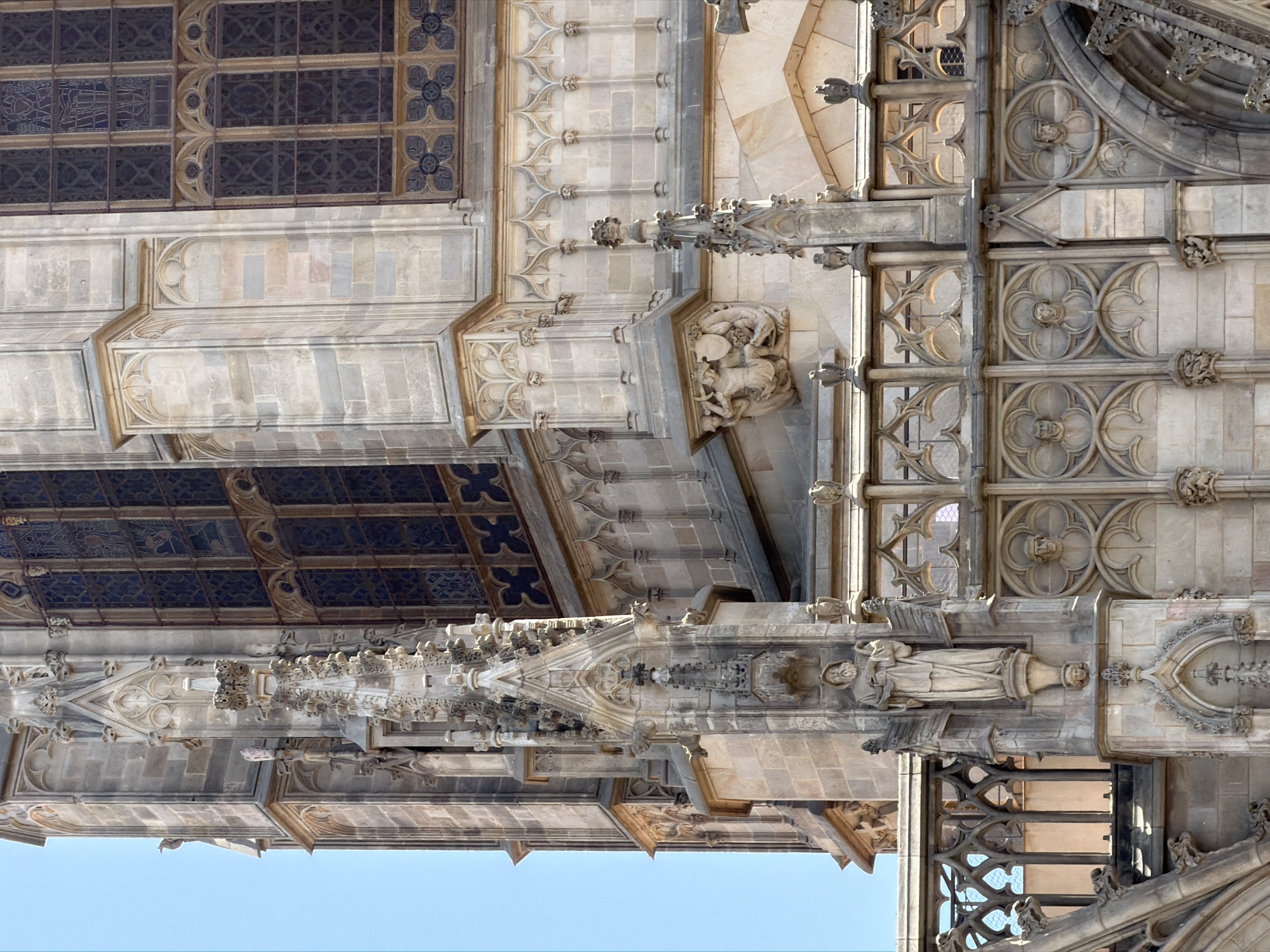 This is a detailed, close-up, exterior photograph of the Barcelona Cathedral, located in the Gothic Quarter of Barcelona, Spain. The image focuses on the intricate stone architecture of the cathedral's facade. Specifically, it captures a section of the upper walls and a prominent spire adorned with gothic-style decorative elements.
Key architectural features include:
- **Stone masonry:** The building is constructed from light-colored stone blocks, exhibiting variations in color and texture, suggesting age and weathering.
- **Gargoyles and sculptures:** Several stone sculptures are visible. One is a statue of a robed figure, possibly a saint or monk, holding a book, positioned in a niche. Another is a relief sculpture depicting a scene with figures, possibly mythological or biblical. Smaller carved heads and decorative elements are also present.
- **Windows:** Large, Gothic-style windows with tracery are visible, some filled with dark glass or stained glass. The windows are tall and narrow, characteristic of Gothic architecture.
- **Spires and pinnacles:** The photograph showcases elaborate spires, pinnacles, and finials, which are pointed decorative elements common in Gothic cathedrals. These are highly detailed with carved ornamentation.
- **Balustrade:** A decorative stone balustrade runs along a lower section of the visible facade, featuring repeating arched patterns and carved medallions or faces.
- **Flying buttresses and supporting structures:** While not fully visible, hints of supporting structures like flying buttresses can be inferred from the architectural complexity.
The sky is clear and blue, indicating a sunny day. The lighting casts shadows that highlight the three-dimensionality and detail of the stone carvings and architectural elements. The perspective is slightly angled upwards, emphasizing the height and grandeur of the cathedral. The image does not contain any people actively engaged in interactions, nor any specific actions. It is a static architectural study.