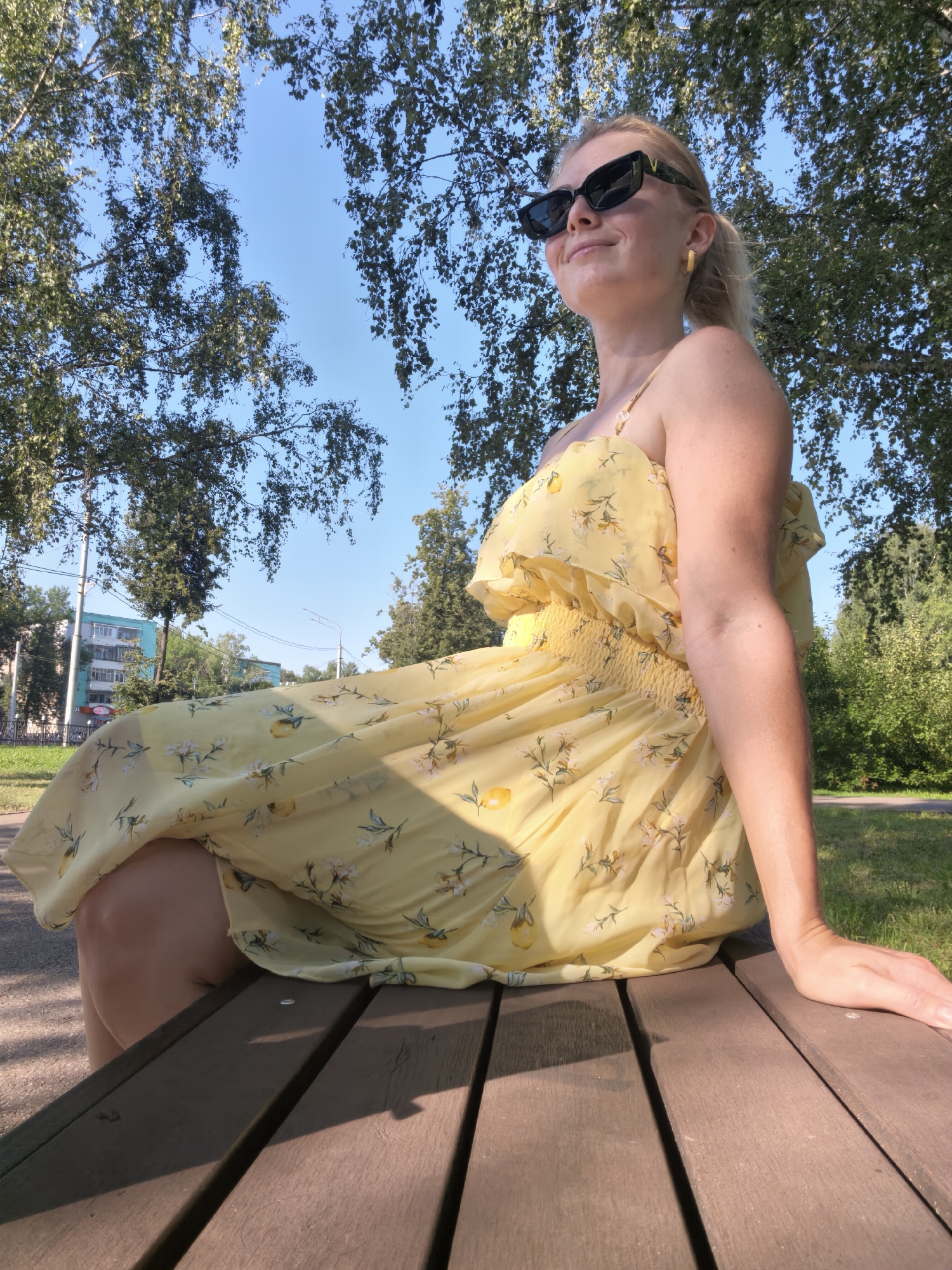 A blonde woman in a yellow floral dress and black sunglasses sits on a wooden park bench. The dress has a ruffled neckline and smocked waist, with a pattern of yellow lemons and white flowers. She is looking to her right with a slight smile. Behind her, trees with green leaves and a clear blue sky are visible. In the background, there are buildings and streetlights, suggesting an urban park setting. The lighting indicates it is daytime, possibly late afternoon due to the shadows cast on the bench. The specific location is a park in the city of Sterlitamak, Russia.