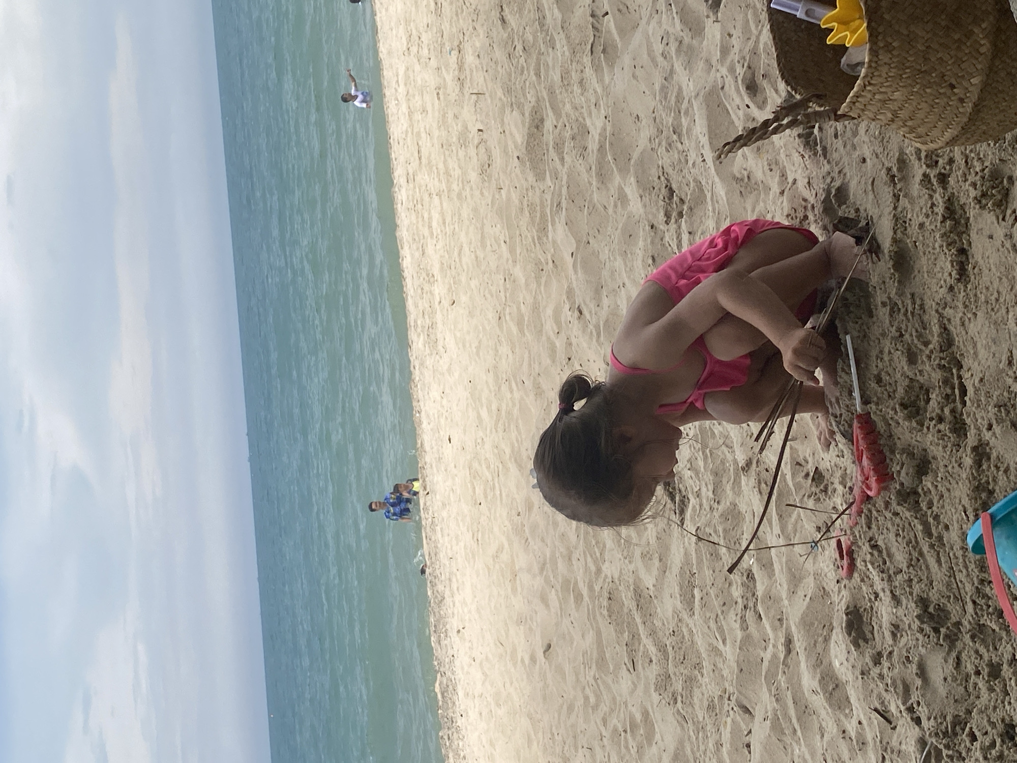 This is a vertical, eye-level, medium shot of a beach scene in Phu Thuy Ward, Vietnam. In the foreground, a young girl in a pink swimsuit is squatting in the sand, playing with sticks. Her back is to the camera, and her hair is pulled up in a ponytail. Beside her are a few sticks and what appears to be a toy shovel. In the background, the ocean stretches out to the horizon under a bright, cloudy sky, suggesting it is daytime. The beach is sandy, with some footprints and rippled patterns in the sand. Further out in the water, a person in a blue and white patterned shirt is visible, wading with two children. Another person in a white shirt is also in the shallow water. The scene conveys a relaxed, family-friendly atmosphere at the beach. The sun appears to be out, but there are clouds in the sky, hinting at potentially variable weather. There is no visible text in the image.