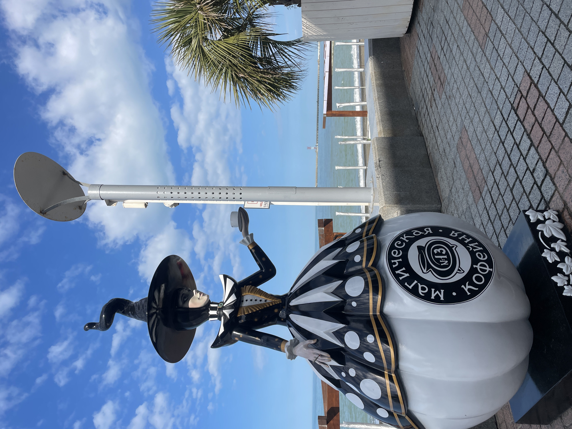 A street-level, wide-angle shot features a quirky sculpture of a woman in a whimsical, black and white outfit and witch hat, next to a modern street lamp, with a palm tree, the sea, and the sky in the background.
The sculpture is the focal point, with a woman in a stylized witch-like attire, primarily in black and white. She's holding a cup in her hand, and her dress is decorated with black and white patterns and golden accents. The base of the sculpture is white with a circular black logo containing text in Russian. A black marble base with white carved floral accents sits next to the figure.
Next to the statue is a gray modern streetlight with a round reflector.
Behind the sculpture, there is a low, white wooden railing with posts, the sea, and a distant structure in the water. The sky above is blue with fluffy white clouds, and a palm tree's fronds protrude from the upper center of the image.
The ground is paved with gray and red rectangular stones, suggesting a pedestrian area or promenade.
The image is likely taken in Sochi, Russia, based on the style of the sculpture and the Cyrillic text.