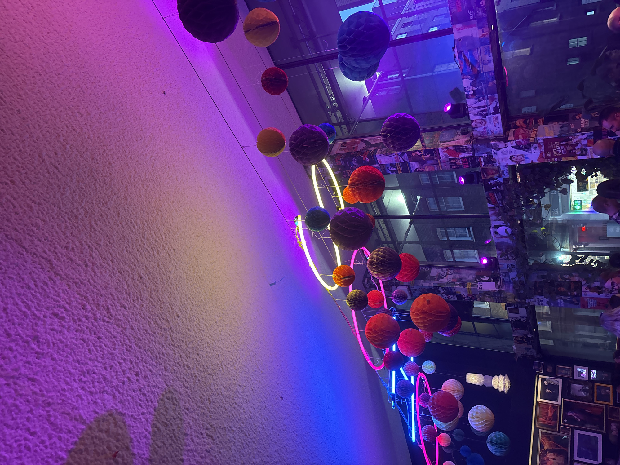 The image captures an interior space in London, United Kingdom, likely a bar or restaurant, decorated with colorful paper lanterns and neon light circles. The lanterns hang from the ceiling, creating a whimsical atmosphere. The background reveals a cityscape reflected in glass windows, featuring architectural details like window frames and building facades. Walls are adorned with an eclectic collection of framed pictures and posters, contributing to the venue's visual texture. Artificial light casts a purple hue over the scene, further enhancing the mood. Patrons can be seen faintly in the reflections and near the edges of the image, suggesting an active environment.
