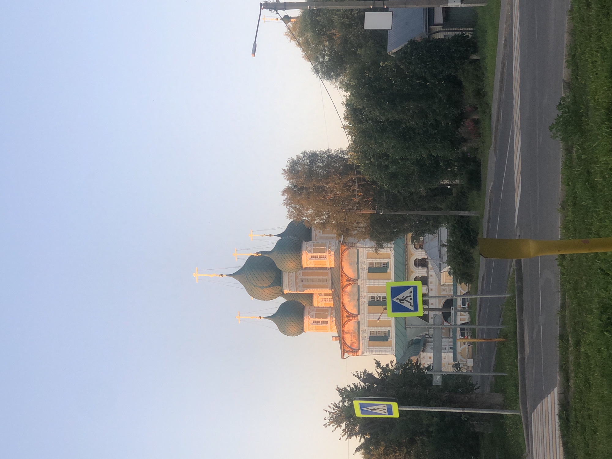 The image shows a view of the Resurrection Cathedral in Tutaev, Russia, captured during the golden hour, likely in the late afternoon or early morning, given the soft, warm light. The cathedral, with its multiple green onion domes topped with golden crosses, is the dominant feature of the scene, showcasing its intricate architectural details. In the foreground, a road with a pedestrian crossing is visible, marked by clear white stripes. Two pedestrian crossing signs are also present, one on the left with a blue background and a white pedestrian silhouette, and another on the right with a light green background and a similar white pedestrian silhouette. Trees line the street and surround the cathedral, adding a natural element to the urban landscape. A street lamp with a lit orange light is visible on the right side of the image. The sky is clear and pale blue, gradually fading to a lighter hue near the horizon. The overall impression is one of serenity and historical grandeur.