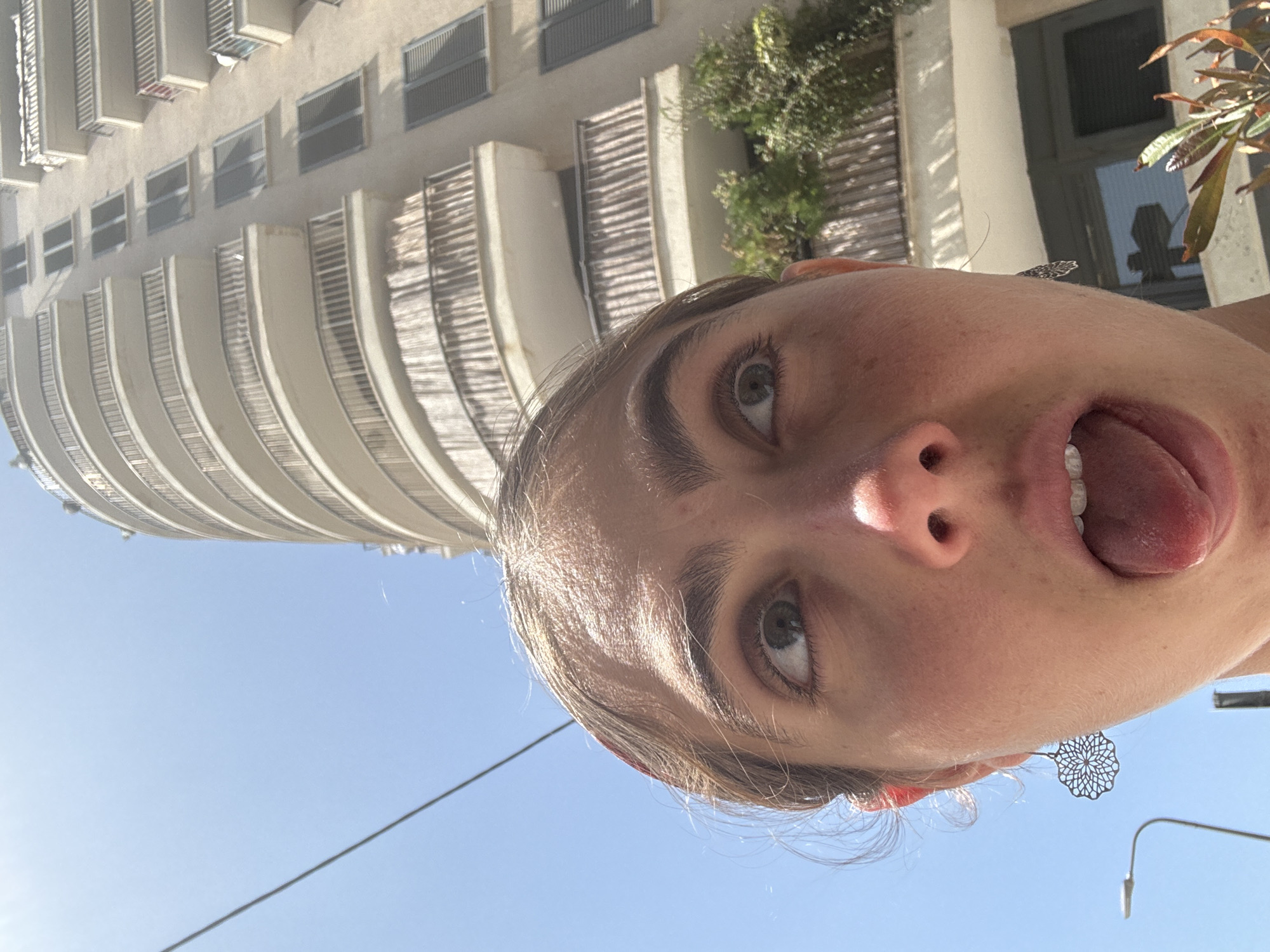 The image is a selfie taken from a low angle, looking up at a woman's face with an open mouth and tongue extended. She has light skin, grey eyes, and brown hair. She is wearing earring. Behind her is a tall, rounded building with multiple balconies and windows. In the upper right corner are some trees and the top of a window or door frame. The sky is blue. Based on the architectural style, the building may be located in Tel-Aviv, Israel.