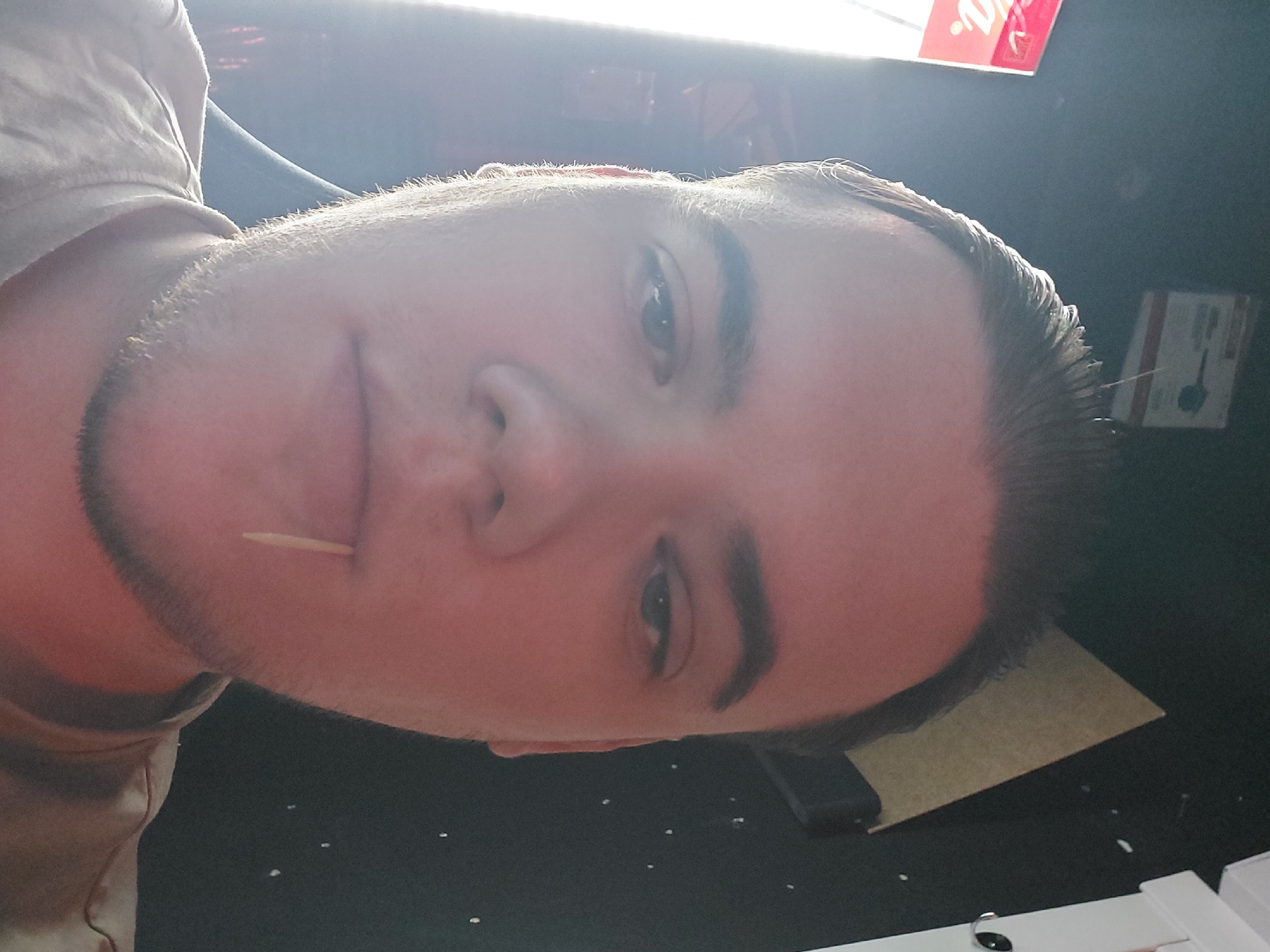 A close-up selfie of a fair-skinned man with dark hair and stubble. He has a toothpick in his mouth. The background is dark but shows hints of a light and possibly a sign with red lettering. There are some office supplies like a brown card and a white box with earphone on the table. There is no way to determine the specific location in Tashkent, Uzbekistan, based on this image alone.