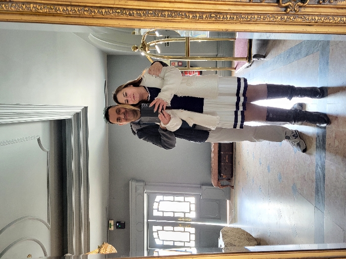 An indoor mirror selfie taken in São Pedro do Sul, Portugal, depicts an adult male and an adult female. The male stands behind the female with his right arm around her shoulder, wearing a dark grey hooded jacket, light-colored trousers, and grey-and-white sneakers, with dark sunglasses resting on his head. The female holds a dark smartphone, directed at the mirror, while dressed in a dark top, a white pleated skirt with two dark horizontal stripes, a white textured cardigan, and dark knee-high boots. Both subjects display slight smiles. The setting features a tiled floor with light and dark geometric patterns, plain light grey walls, and a white ceiling adorned with multiple decorative moldings. Reflected in the background are light-colored double doors, a large stone object, an upholstered bench positioned above a dark trunk, a gold-colored luggage cart, and a red fire extinguisher cabinet. A chandelier is partially visible in the upper left reflection.