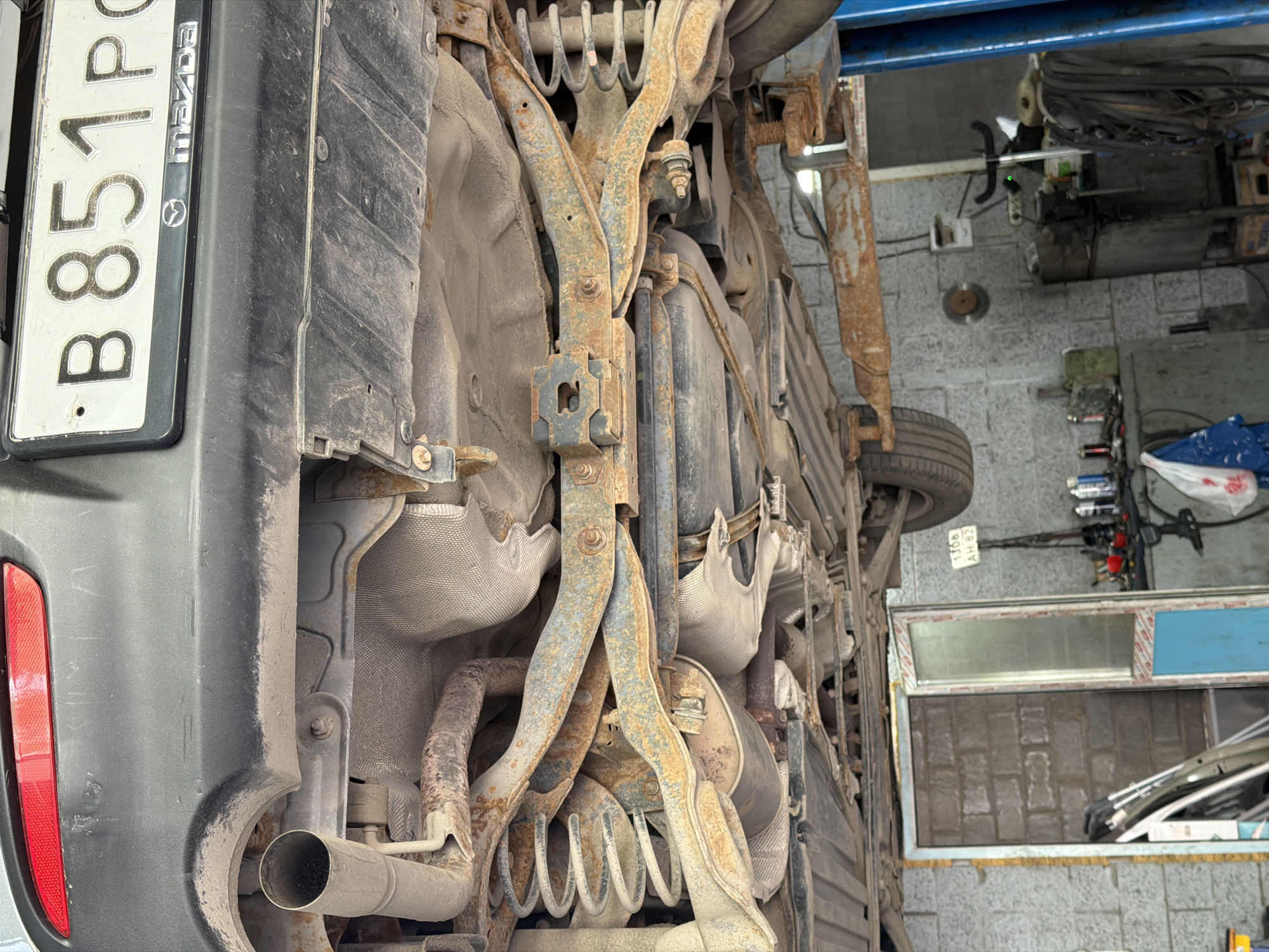 The image shows the underside of a car that is elevated on a lift, likely in a repair shop. The car's exhaust system and suspension components are visible, with significant signs of rust on many of these parts. The license plate of the car reads "B 851 PC" and has a Mazda logo. In the background, a brick wall and various tools and equipment are present, suggesting an industrial or workshop setting. A sign with Cyrillic text, "1308 AH 82", is visible hanging on the wall. This scene is consistent with a location in the city of Ягул, Russia.