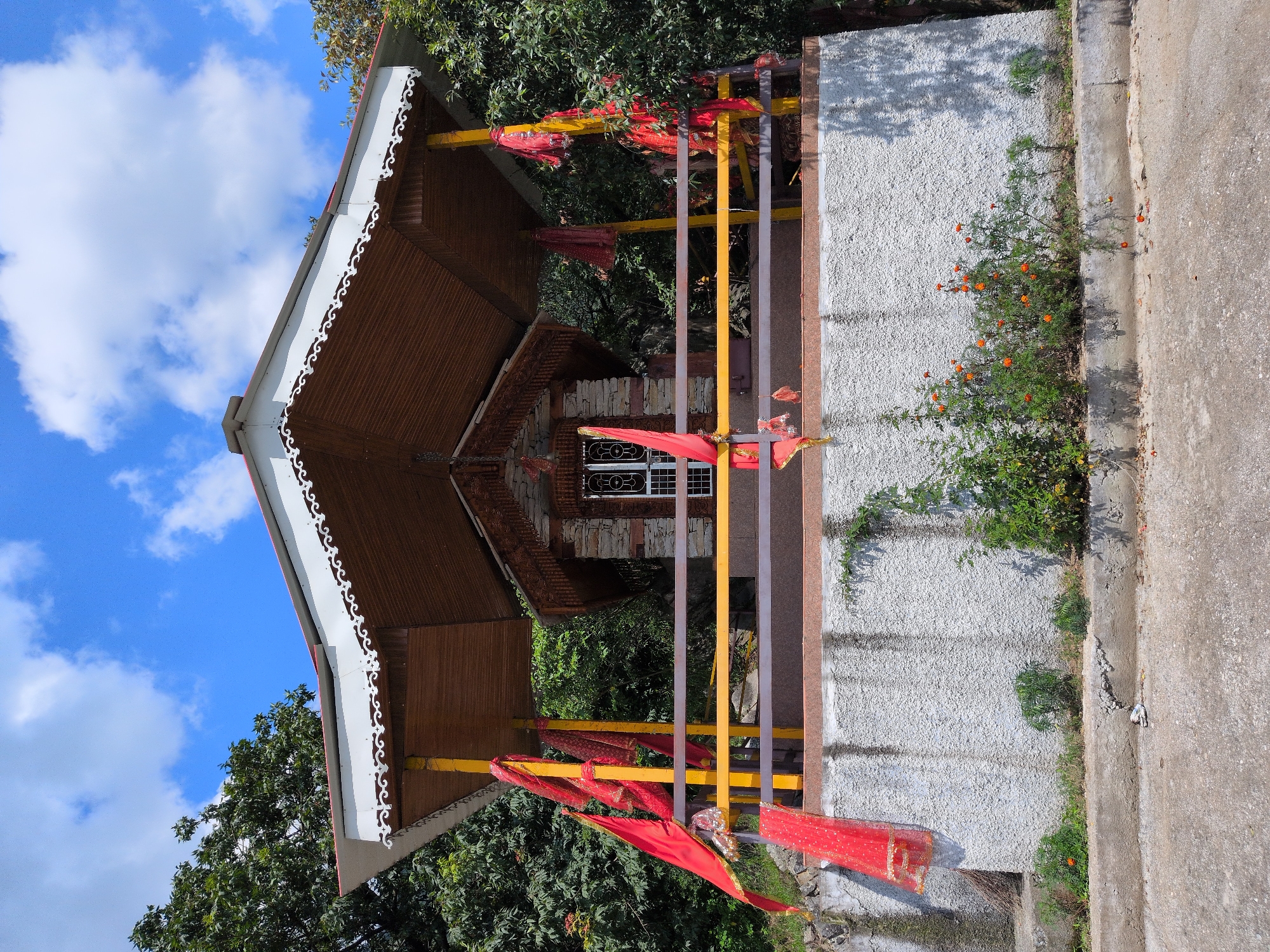 In Bagicha, India, a small, open-air shrine with a wooden roof and stone walls stands against a backdrop of lush green trees and a bright blue sky with scattered clouds. The shrine is decorated with red fabric draped over yellow pillars and railings, suggesting a religious or cultural significance. Orange flowers bloom at the base of the shrine, adding a touch of color to the scene. The shrine is situated on a raised platform with a concrete base, and a paved pathway leads to it. The overall impression is one of tranquility and natural beauty.