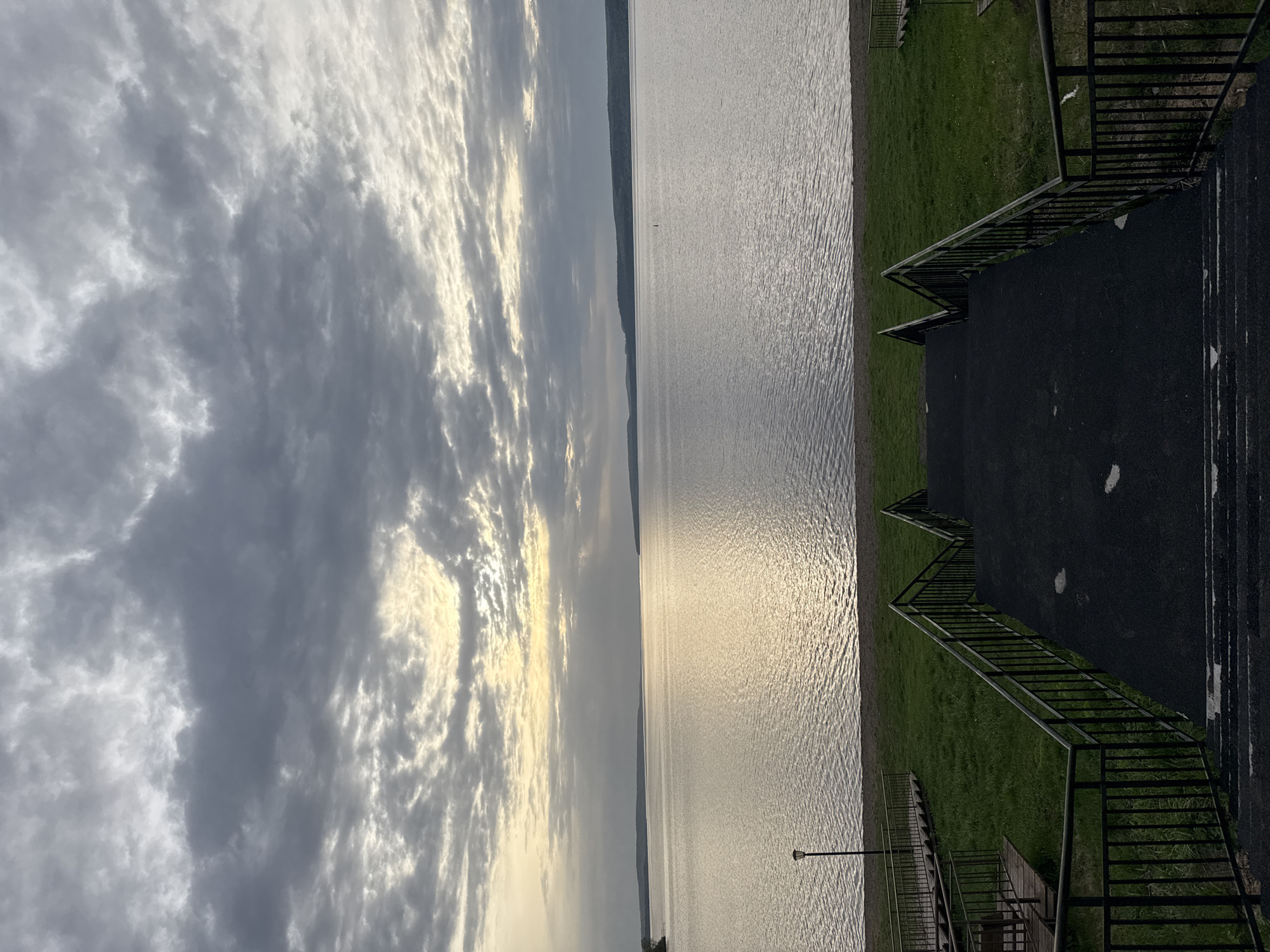 The image shows a body of water under a cloudy sky with sunlight breaking through. A strip of land with green grass runs along the water's edge, with a black paved path and metal railings visible. These railings form steps going from the lower level by the water, up to a higher pavement. A light post can be seen at the end of one staircase. The location is Osa, Russia.