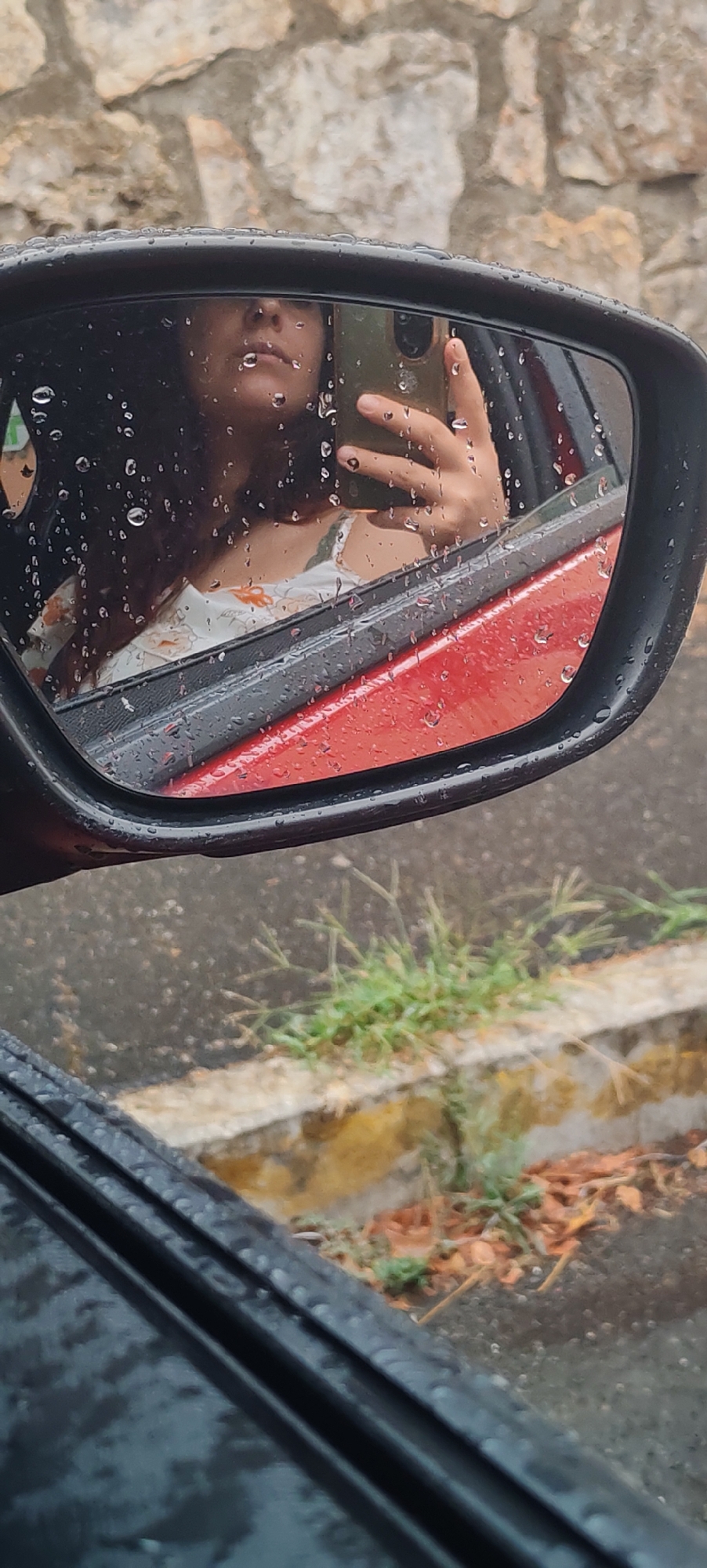 The image is a close-up, partially obscured reflection in a car's side mirror on a rainy day. The mirror itself is framed by a black casing and shows numerous water droplets on its surface.
Reflected in the mirror is a person's face, specifically the lower half of their face, including their nose and mouth. The person is holding a smartphone in their right hand, presumably to take the selfie. The phone is a light green color. The person appears to be wearing a white dress with an orange floral pattern. Their dark hair is visible, falling around their face. The reflection also shows the interior of a red car.
The background visible in the mirror is a textured stone wall. Outside the mirror's reflection, the foreground shows a wet asphalt surface with patches of green grass and some dry leaves. A concrete curb or ledge is also visible. The overall environment suggests an outdoor setting, likely a street or roadside in a city, given the presence of a car, asphalt, and urban-style stone construction. The presence of water droplets indicates it is raining or has recently rained.
There is no information within the image to suggest the location is the city of La Spezia, Italy.