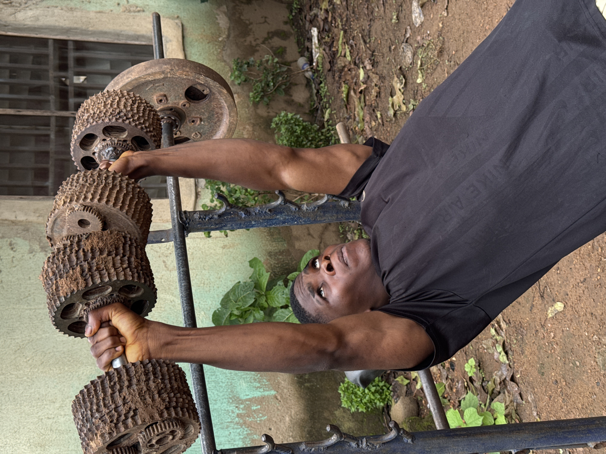 A Black man is performing a bench press exercise with a homemade barbell. The barbell is constructed from rusty gears and metal plates, possibly repurposed industrial waste. The man is lying on a rudimentary bench press rack made of welded metal, painted blue. He is wearing a black t-shirt with the "NIKE AIR" logo subtly visible. The background consists of an outdoor environment, likely a yard or open space, with patchy greenery, fallen leaves, and a wall with a window. The overall impression is one of an improvised gym setup in a potentially less developed urban or semi-urban area, consistent with the context of Akwanga, Nigeria.