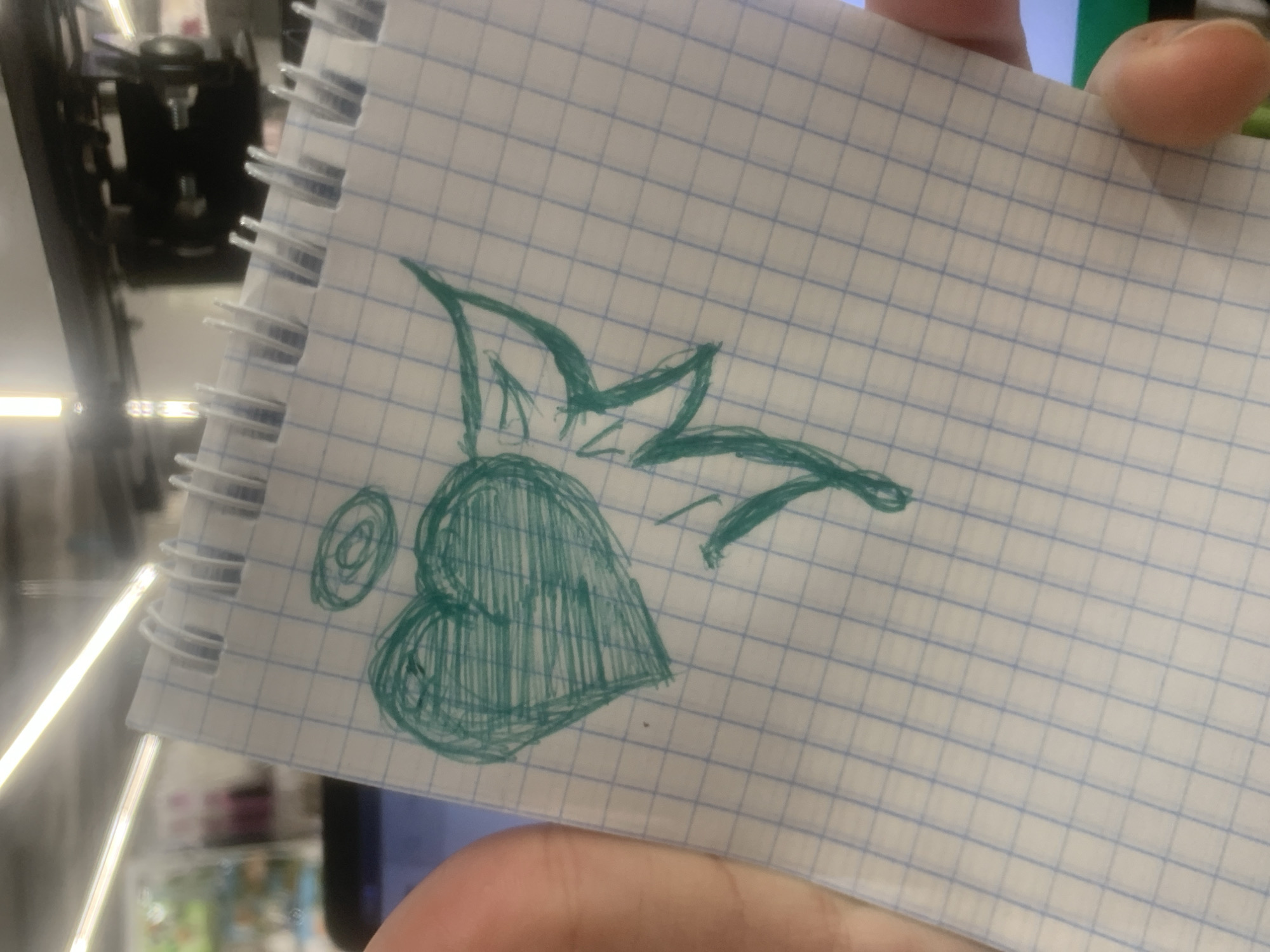 An overhead, close-up shot shows a person's left hand holding a squared notebook open. A turquoise, hand-drawn sketch of a heart with a halo and wings is centered on the notebook page. The notebook is spiral-bound and has blue grid lines. The background is out of focus, showing a blurred room interior with fluorescent lighting and what appears to be a shelf or structure in the upper portion of the image.