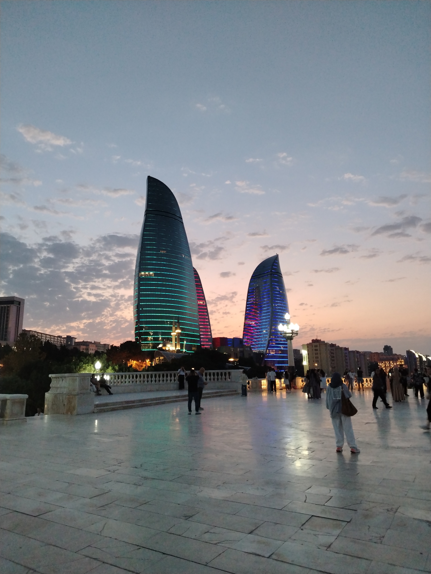 The image depicts the Flame Towers in Sabail Raion, Azerbaijan, at dusk. The distinctive flame-shaped skyscrapers are illuminated with vibrant blue and pink lights, creating a striking contrast against the fading twilight sky filled with scattered clouds. In the foreground, a paved plaza with a marble-like surface reflects the city lights. A white ornamental balustrade separates the plaza from a lower level where several individuals are seated or standing. Other people are seen walking on the plaza, some in casual attire, including one woman in a headscarf. The overall scene captures a moment of urban beauty and activity as day transitions into night in a prominent architectural landmark of Baku.