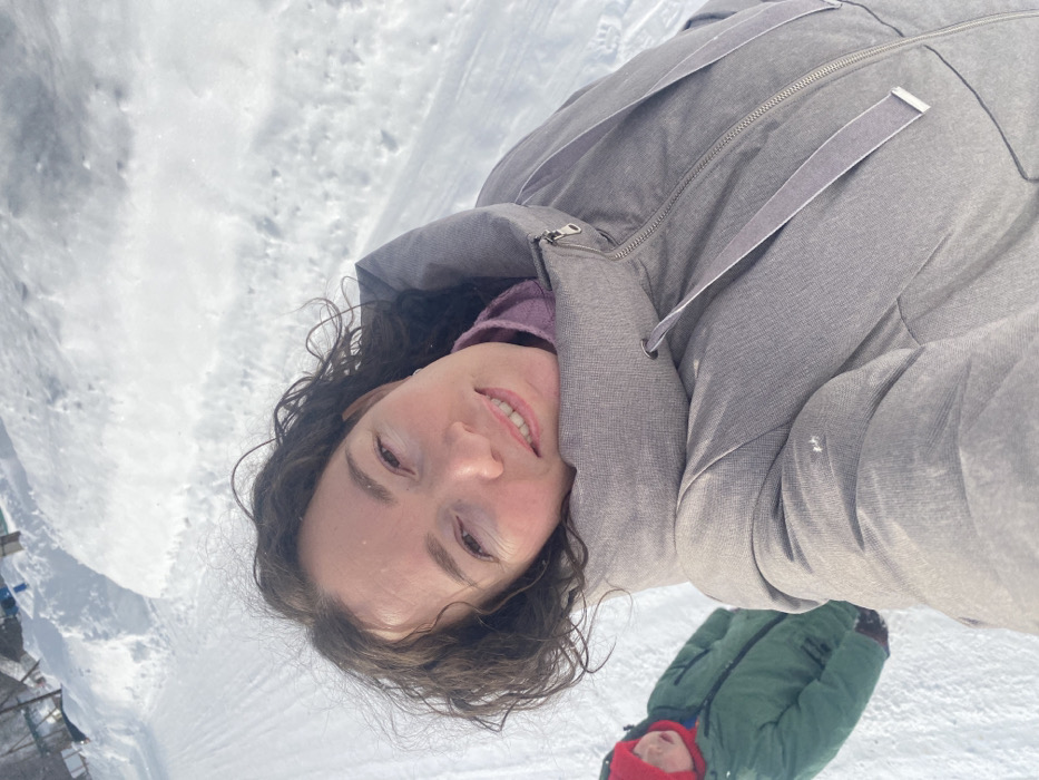 The image depicts a winter outdoor scene in the city of Узяр, Russia. In the foreground, a woman with dark, curly hair, wearing a grey puffer jacket over a purple garment, smiles directly at the camera in a selfie-style shot. To her left and slightly behind her, a child wearing a red hat and a green winter jacket is partially visible, with their mouth open. The ground is entirely covered in snow, with a track or path visible in the middle distance. High snowbanks line the sides of this path. The environment appears bright, indicating daylight conditions.