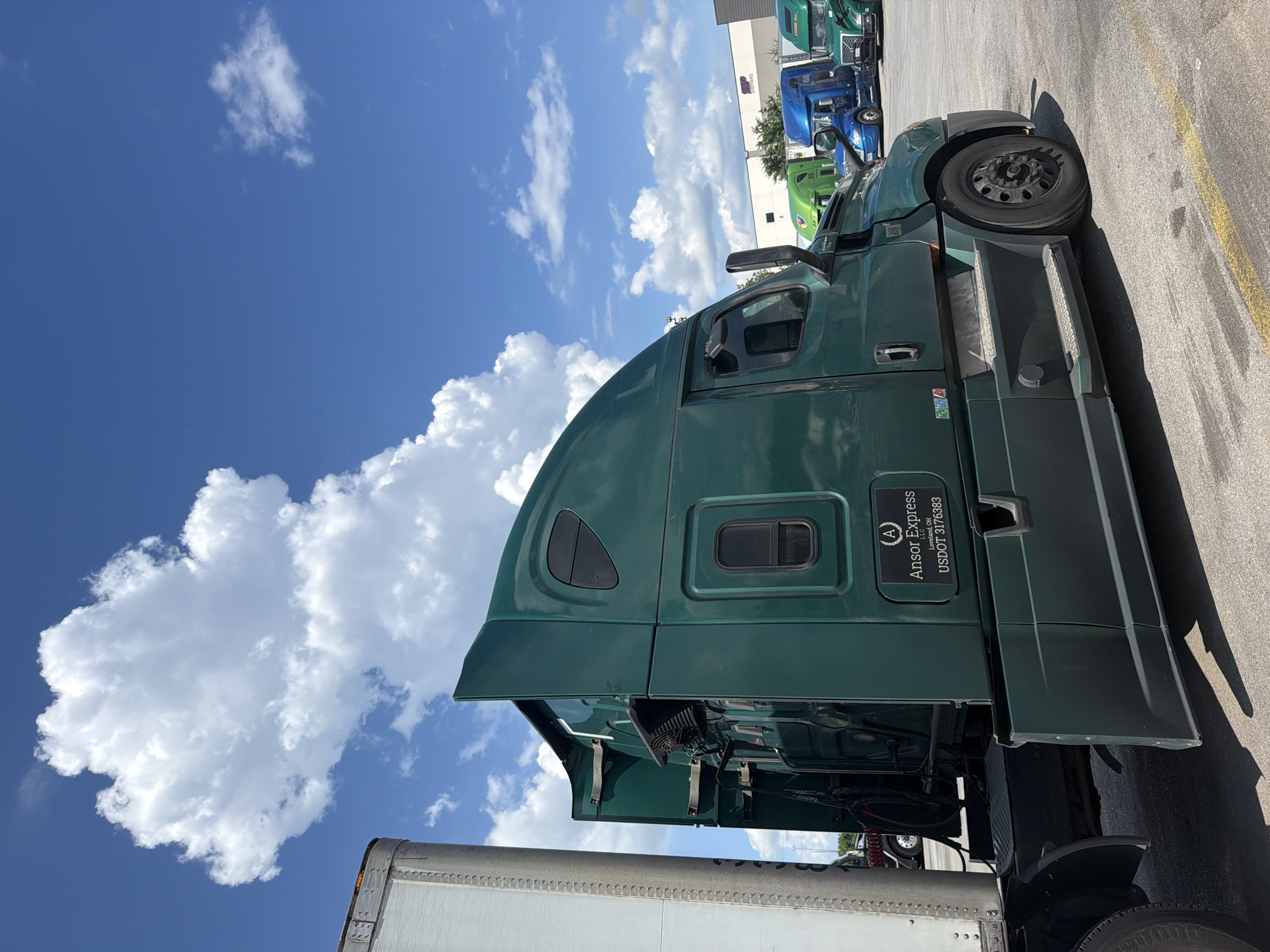 The image shows a row of semi-trucks parked in an outdoor lot under a partly cloudy blue sky. The main focus is on a dark green semi-truck in the foreground, seen from its right side. This truck has the logo "Ansor Express LLC" with "Loveland, OH" and "USDOT 3176383" printed on a black rectangular decal on its side. A portion of a white trailer is visible to the left of the green truck. In the background, other semi-trucks of various colors, including blue and bright green, are parked. A light-colored building with trees in front of it is visible in the far background. The ground is asphalt with yellow painted lines. There are no people visible in the image. The location is the city of Lebanon, United States of America.