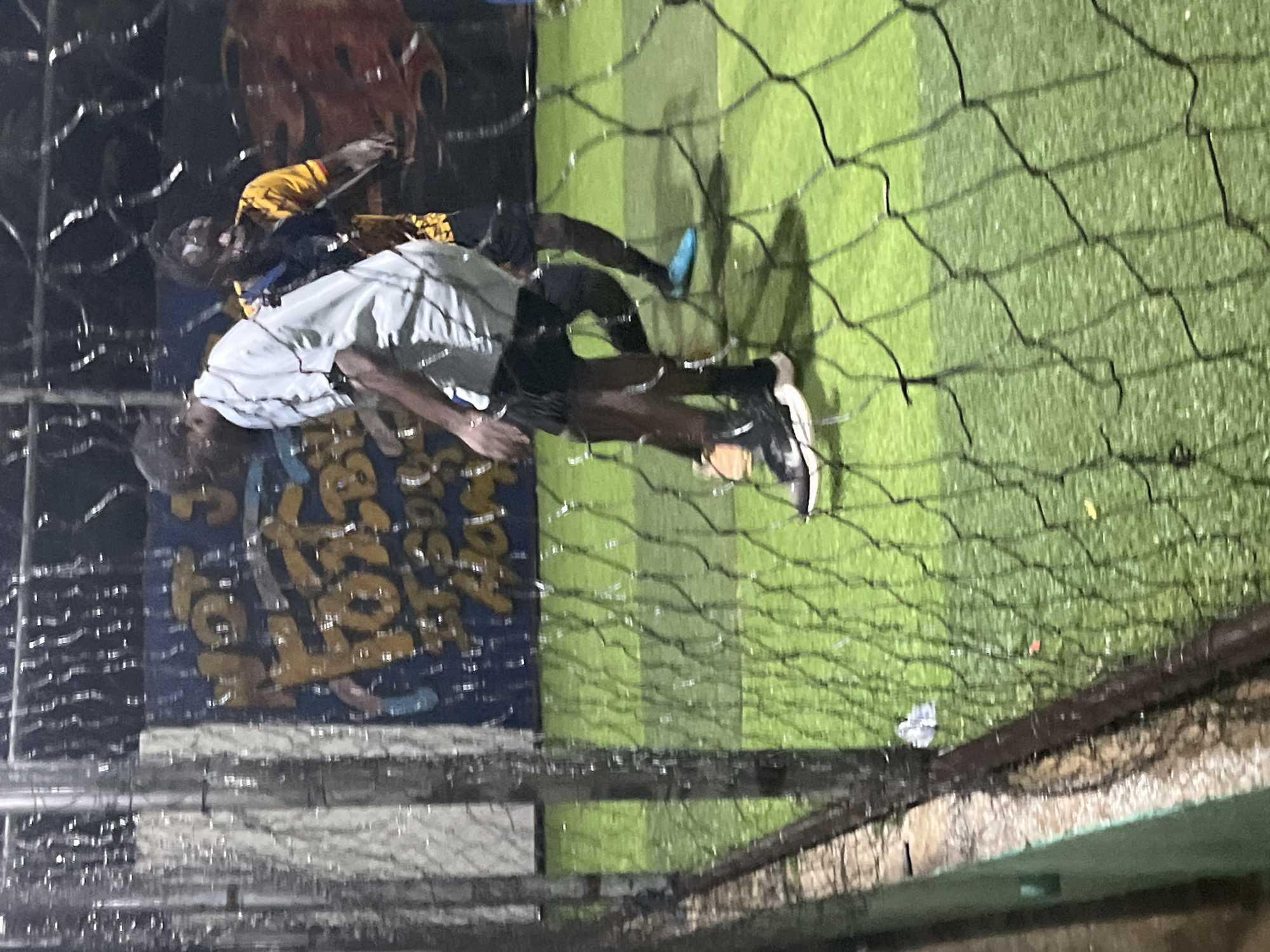 The image depicts a nighttime scene of two individuals playing soccer on an artificial turf field, likely in Jos, Nigeria. The field is enclosed by a fence, part of which is visible in the foreground, covered by a net that partially obstructs the view. The ground is a vibrant green, and the lighting suggests it is an outdoor facility illuminated for night play.
One player, wearing a light-colored short-sleeved shirt and dark shorts, is in motion, with his right foot near a soccer ball. His shoes are dark with white soles. The second player, to the right and slightly behind, is wearing a yellow and black patterned shirt and dark shorts, and appears to be reacting to the ball or the other player's movement. He has blue cleats on his feet.
In the background, a colorful mural or advertisement is visible on a wall, featuring yellow and blue lettering that is partially obscured by reflections and the netting. The overall impression is of a casual, perhaps amateur, soccer game taking place under artificial lights. The presence of the net and the enclosed nature of the field suggest it is a dedicated sports facility.