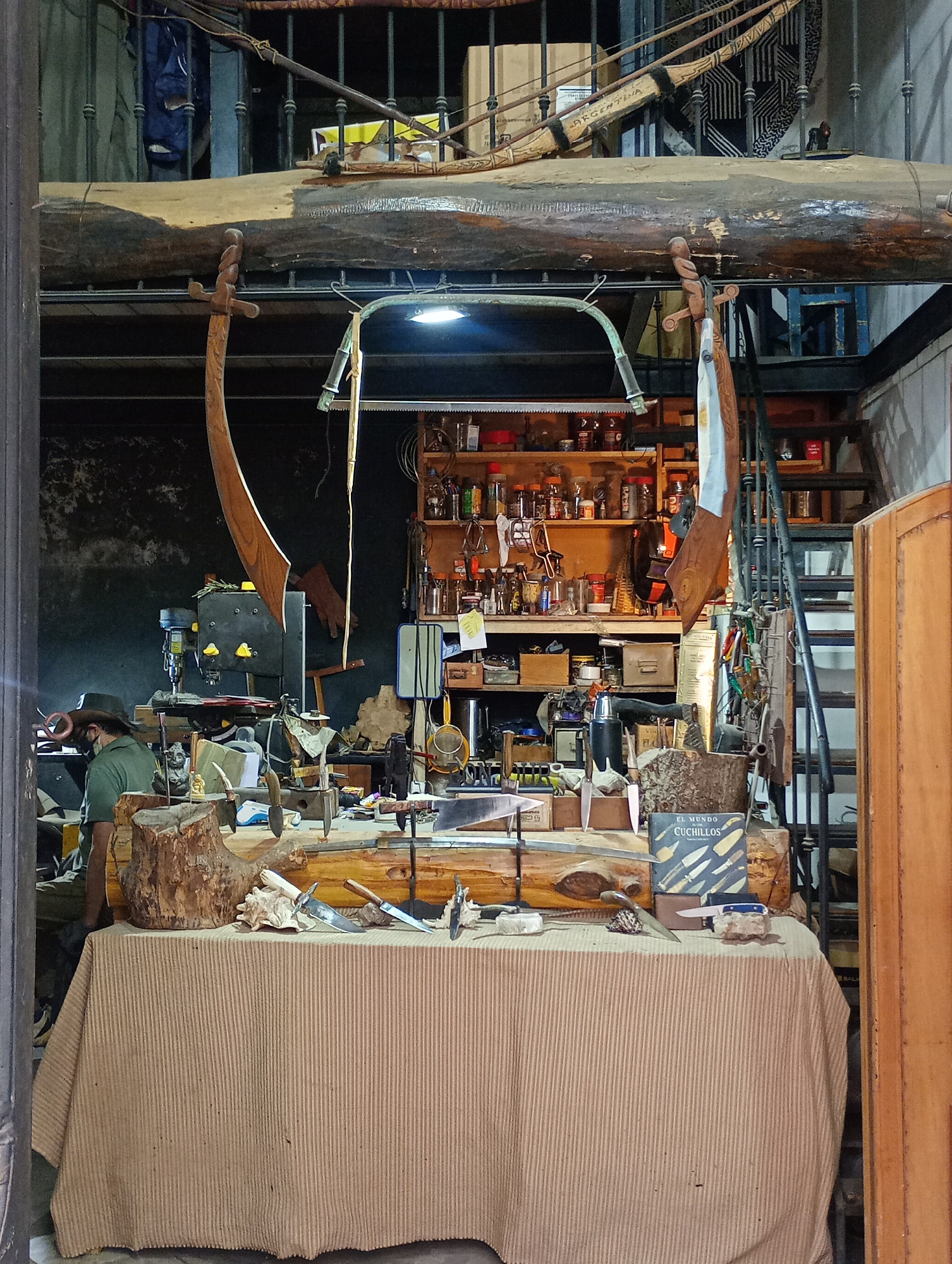 The image shows the interior of a workshop or studio, potentially in Buenos Aires, Argentina. A man wearing a hat and a face mask is visible on the left side of the frame, standing near a cluttered workbench. The workbench is covered with a striped tablecloth and holds various items, including knives, pieces of wood, a seashell, and a book titled "EL MUNDO CUCHILLOS." Behind the workbench, a cluttered shelving unit is filled with jars, tools, and other objects. Stairs are visible on the right, leading to an upper level. Above the workbench, a large log is suspended horizontally. A wooden sword hangs from it, and a bow labelled "ARGENTINA" sits atop. The overall impression is of a busy, creative space, possibly belonging to a craftsman or artisan.