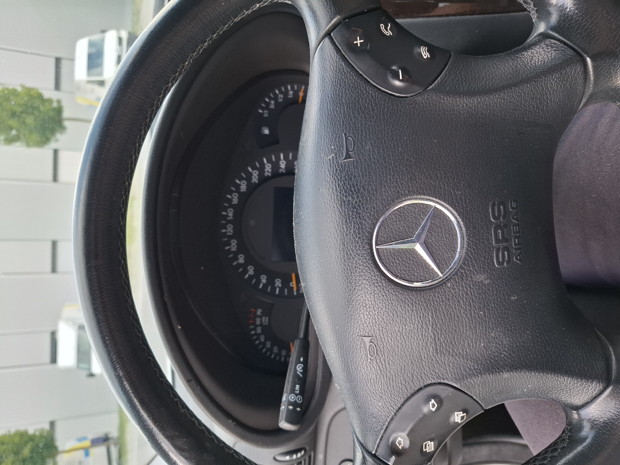 The image shows the interior of a Mercedes-Benz vehicle, specifically the steering wheel and dashboard from the driver's perspective. The steering wheel is black leather with a silver Mercedes-Benz emblem in the center. Control buttons are visible on the left and right sides of the steering wheel. The dashboard includes a speedometer and a fuel gauge. A turn signal stalk with cruise control buttons is visible on the left side of the steering column. Outside the vehicle, two white vans and trees are visible through the windshield, suggesting an outdoor setting. There is no visible indication of the location being in Rothenburg, Switzerland, nor are there any people or specific actions depicted.