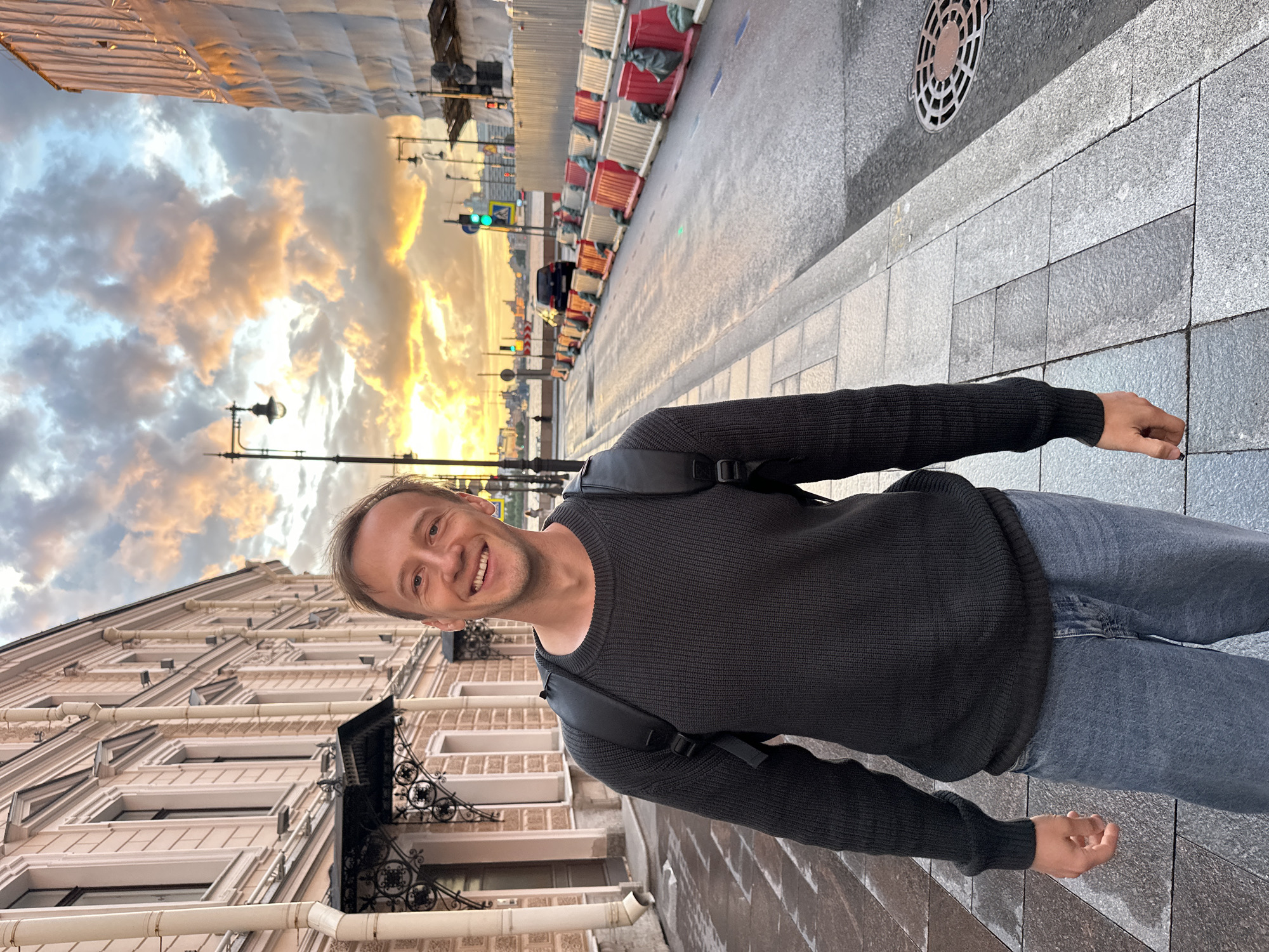 A man is standing on a sidewalk in Saint Petersburg, Russia, facing the camera with a smile. He is wearing a dark knitted sweater and blue jeans, and a black backpack. The sidewalk is made of stone tiles. Behind him, to his left, is a building with light-colored facade and ornate windows. To his right, the street is lined with construction barriers and a car is driving down the wet road. The sky is dramatic with colorful clouds illuminated by the setting sun. A lamppost and traffic lights are visible in the distance. The overall scene suggests an urban setting during the golden hour.