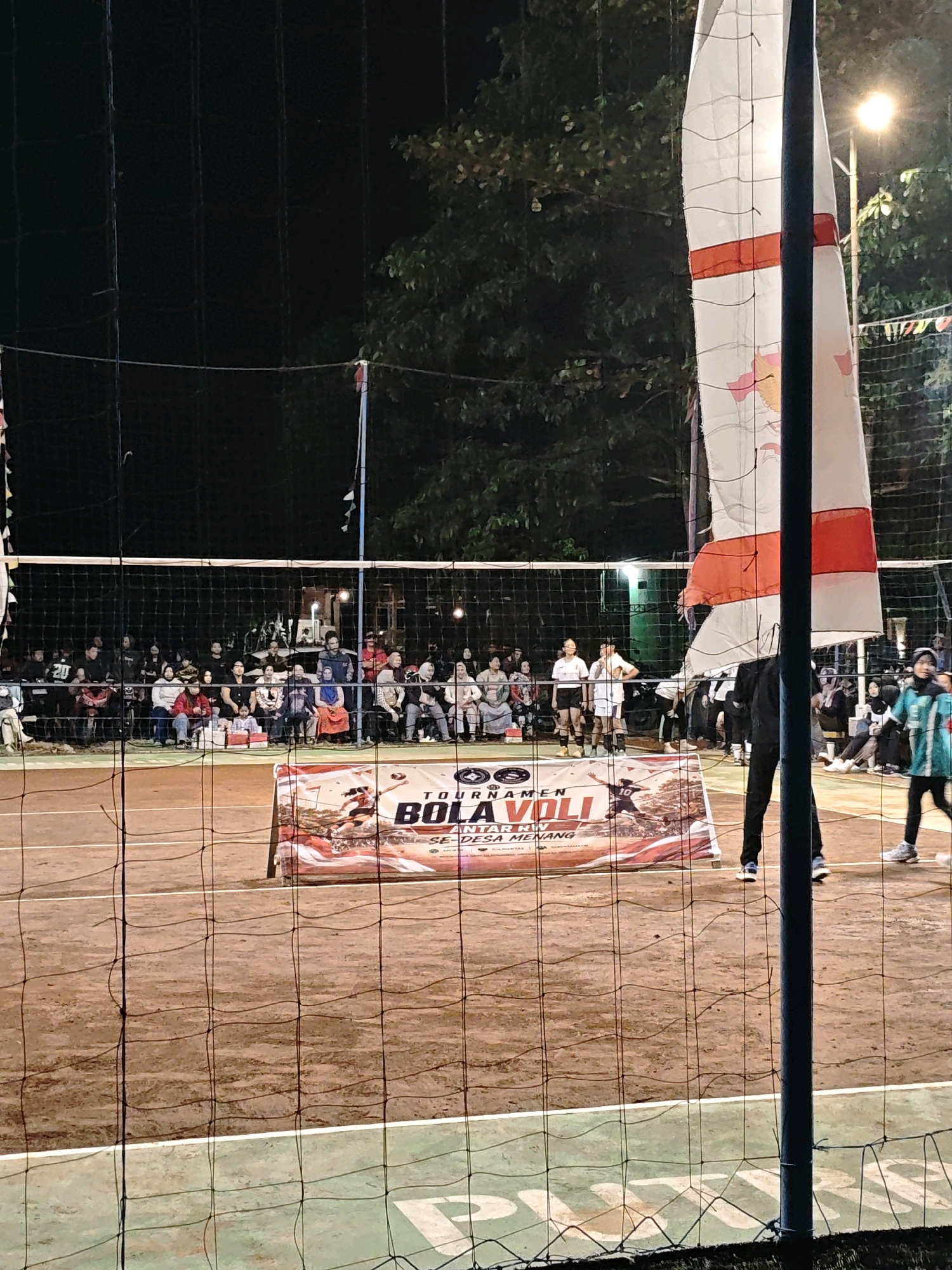 An outdoor volleyball tournament is in progress at night on a dirt court in Pagu, Indonesia. Spectators are seated in bleachers behind a net. A large banner is displayed on the court, featuring images of volleyball players and text in Indonesian. A tall flagpole with a white and red flag stands to the right of the frame. Several floodlights illuminate the court.