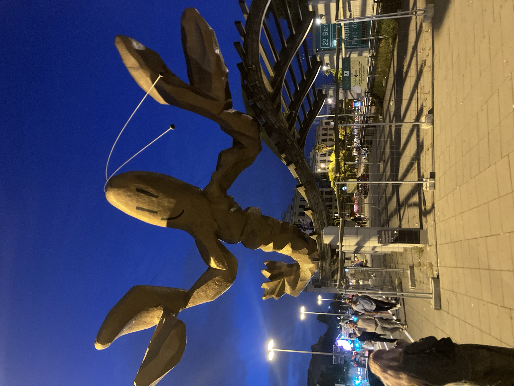 The image captures an outdoor scene at dusk in Barcelona, Spain, dominated by a large, golden sculpture of a lobster or crayfish. The sculpture is depicted in a dynamic, leaping pose, with its claws extended. It features a simple, smiling face with two black lines for eyes and a curved line for a mouth, and thin white antennae extending from its head. The sculpture is mounted on a dark, curved, trellis-like structure.
In the foreground, a woman with long, blonde hair is seen from behind, facing away from the camera and towards the sculpture. She is wearing a dark jacket and carrying a dark bag over her shoulder. The ground she stands on is paved with square, light-colored tiles. Beyond her, several people are walking on a sidewalk, some in casual attire. The area appears to be under some form of construction or renovation, as temporary metal fencing with concrete bases encloses parts of the scene.
In the background, streetlights illuminate the area, casting long shadows. Signs are visible, including a green directional sign indicating "22 Barb." and "Hospital Parc de..." suggesting proximity to a hospital and a park. Buildings with illuminated windows are visible behind the fencing and vegetation. The sky is a deep blue, transitioning into twilight. The overall impression is of a public space in a city during the evening hours, featuring a prominent art installation.