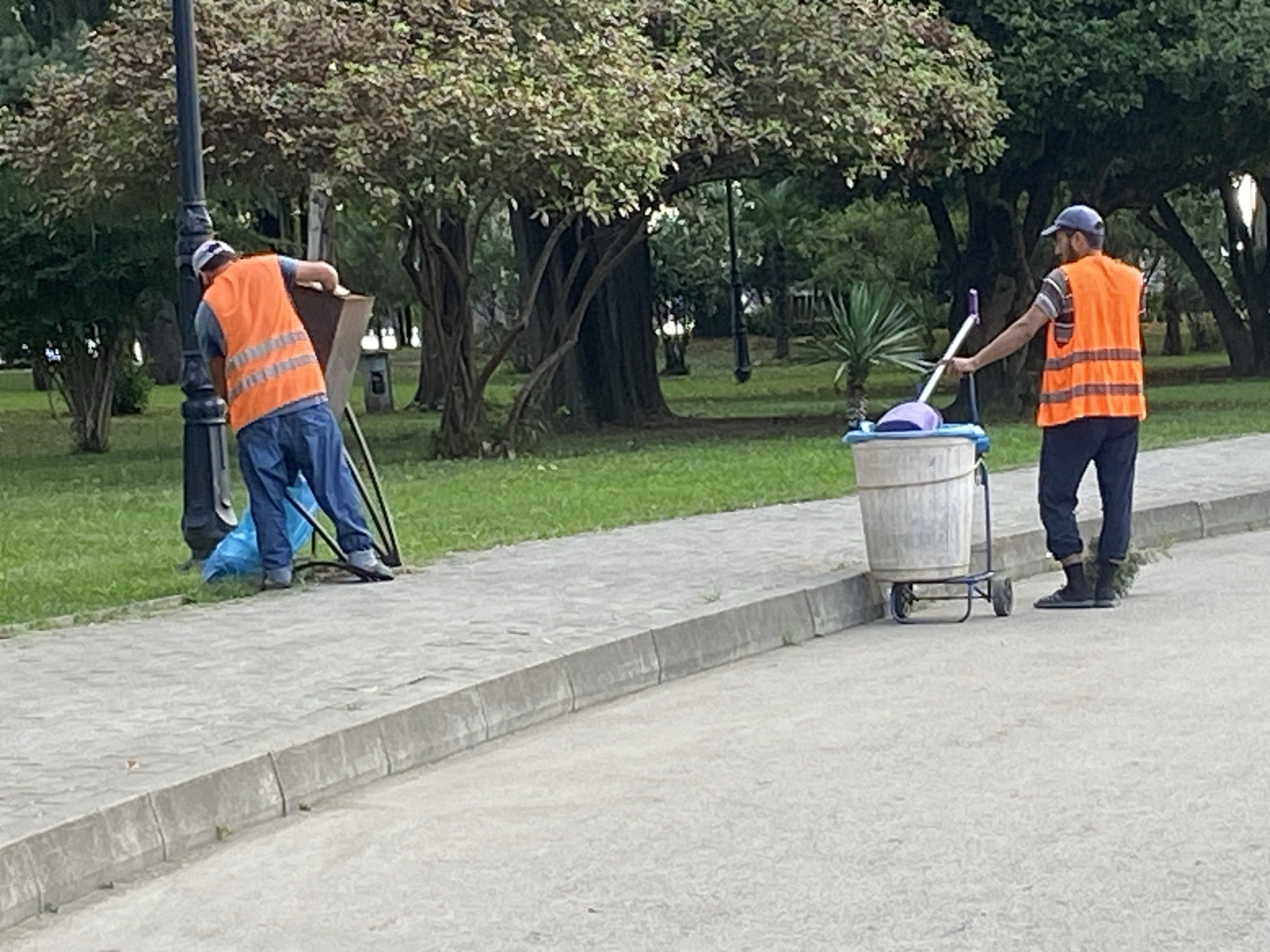 Two sanitation workers are visible on a paved walkway in a park-like setting. The worker on the left, wearing a white cap, orange reflective vest, and blue jeans, is bent over, attending to a trash receptacle attached to a lamppost. A blue trash bag is visible on the ground next to the receptacle. The worker on the right, wearing a blue cap, orange reflective vest, and dark pants, is pushing a cart with a large bin and a broom. The background consists of lush green grass and mature trees, with a lamppost visible in the distance. The scene appears to be in an outdoor public space in the city of Sokhumi, Abkhazia.