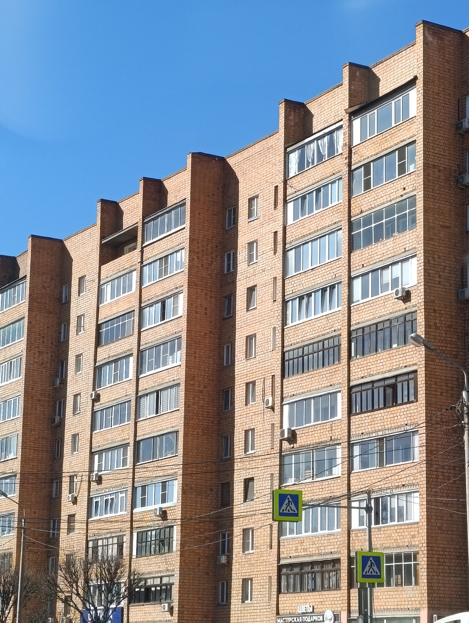 The image shows an orange brick apartment building in Krasnoyarsk, Russia, under a clear blue sky. The building has multiple floors with numerous windows, some of which have air conditioning units attached. In the foreground, there are pedestrian crossing signs. Street lights and power lines are also visible. Bare trees are present at the base of the building. A sign with Cyrillic writing is partially visible at the bottom of the image.