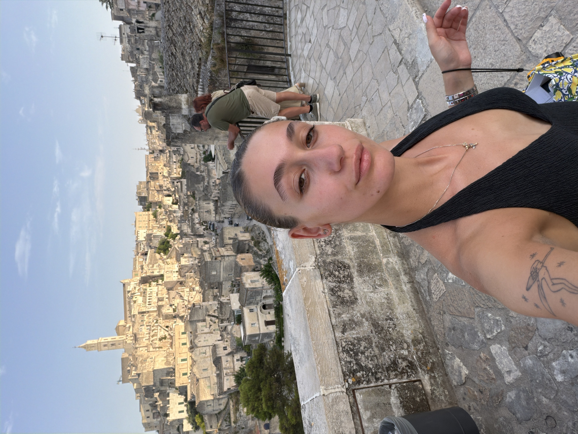 In a selfie taken in Matera, Italy, a young woman with her hair pulled back is centered in the foreground, her face turned slightly toward the camera. She is wearing a black halter top, a necklace with a small pendant, and a bracelet on her left wrist. Her right arm is visible, displaying a line-art tattoo of a figure with wings and small stars.
Behind her, a couple stands by a railing, looking out at the Sassi of Matera, a UNESCO World Heritage site renowned for its ancient cave dwellings and historic architecture. The woman in the couple has red hair and wears a light-colored dress, while the man is in a green t-shirt and beige shorts. The background depicts the characteristic honey-colored stone buildings cascading down a hillside under a clear, pale blue sky. The ground in the foreground and middle ground is paved with stone tiles. A trash can is visible to the left of the woman.
