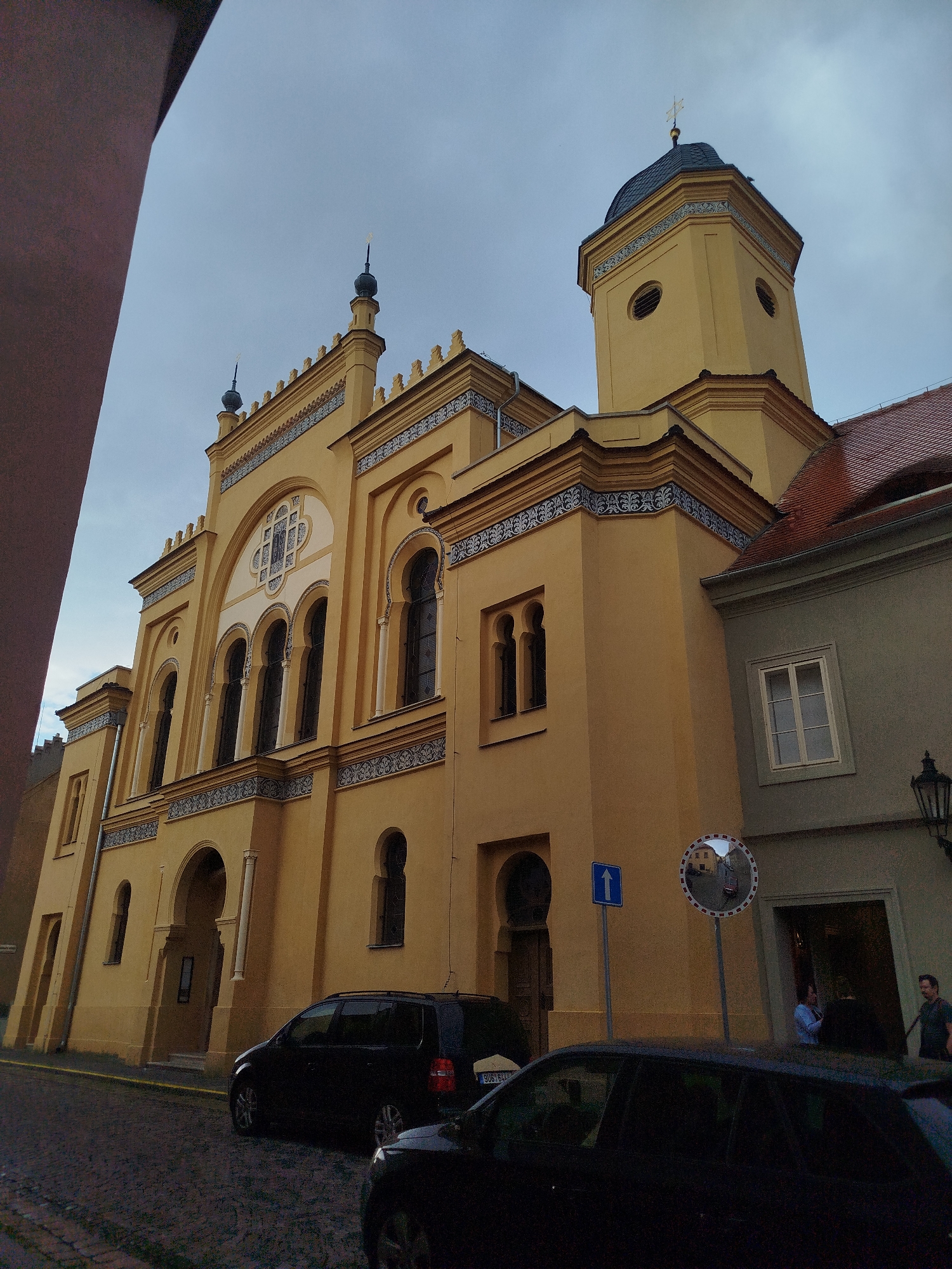 The image depicts a view of a yellow building with ornate architectural details, likely a synagogue, under an overcast sky. The building features multiple arched windows, decorative friezes, and a prominent tower with a dark roof and a cross at its apex. To the left of the main building, a dark structure partially obscures the frame. In the foreground, a cobblestone street is visible with two dark-colored cars parked. One car is a black SUV, and another is a dark sedan. A street sign with an upward-pointing arrow and a circular mirror with a reflection of buildings and a street are positioned near the cars. A lamppost is visible on the right side of the frame. Two individuals are seen standing near the entrance of an adjacent building on the right. The overall scene suggests an urban environment, possibly in a historic district.