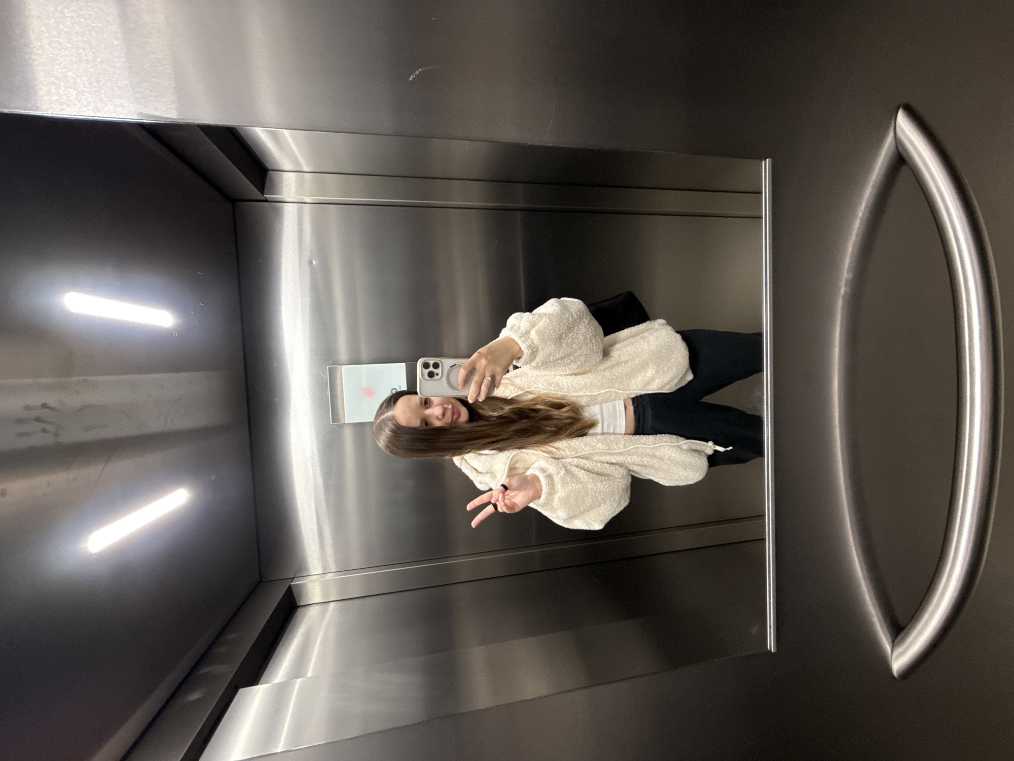 A woman stands in an elevator, taking a selfie in the mirror. She is holding a phone and making a peace sign with her other hand. She wears a fluffy, off-white jacket, a white top, and dark pants. The elevator interior is metallic with two horizontal light fixtures on the left. A curved handle is attached to the elevator door on the right. The location is unknown and there is no information to suggest it is in Barcelona, Spain.