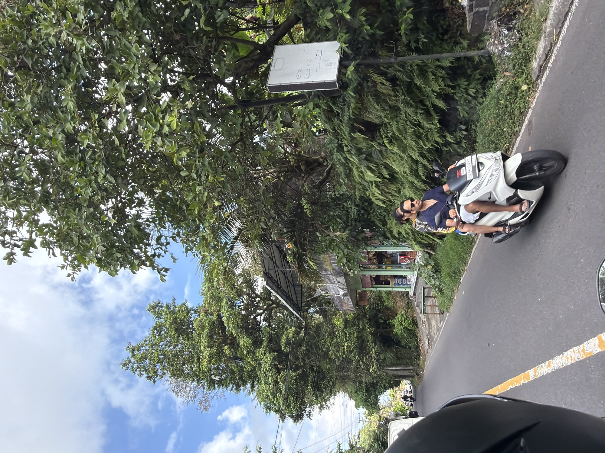 The image captures a scene on a road in Tampaksiring, Indonesia. The foreground shows the helmet of a rider, indicating a first-person perspective from a motorcycle. To the right, a white scooter is seen with two individuals riding on it. The male rider in front is wearing a navy blue shirt and white shorts, with sunglasses on. The female passenger behind him is wearing a patterned dress. They are moving along the asphalt road which has a broken yellow line marking the lane. The environment is lush and green, with tall trees and dense foliage lining the sides of the road. A small building with a storefront, possibly a shop, is visible on the right side of the road, nestled among the greenery. A white sign on a pole is also present. In the distance, the road continues, with other vehicles and more trees visible. The sky is blue with scattered white clouds.