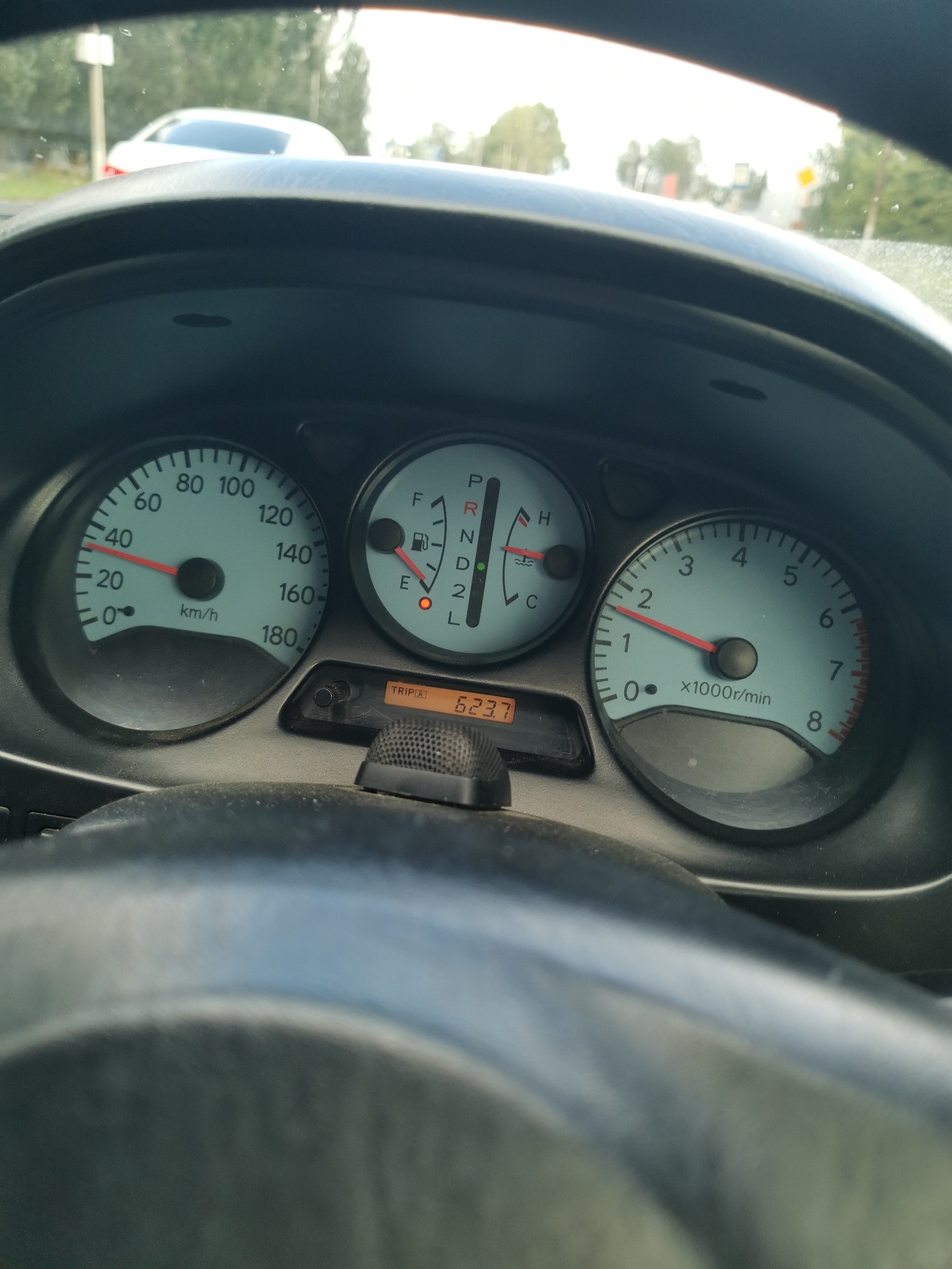 This is a close-up, first-person perspective image taken from inside a car, focusing on the dashboard and steering wheel. The dashboard features a speedometer, a tachometer, a fuel gauge, and a gear indicator. The speedometer shows a speed of approximately 40 km/h. The gear indicator displays "L" for low gear, and the fuel gauge is near empty. A digital display shows "TRIP A 623.7". The scene appears to be set during the daytime, with natural light illuminating the interior of the car. Through the windshield, a blurred background shows trees and a road, suggesting the car is in motion in an outdoor setting. There are no people visible in the frame, and the focus is entirely on the car's controls. The overall impression is one of a routine drive, with the driver capturing a snapshot of their car's dashboard. The location context provided is Oktyabrsky, Russia.