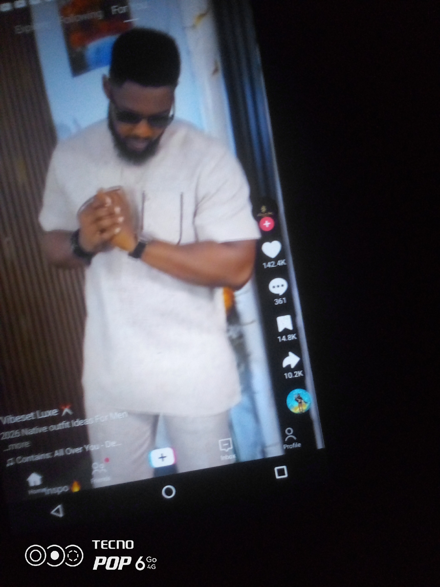 The image displays a close-up of a smartphone screen, likely a TikTok interface, showing a video of a man. The man is wearing a light-colored traditional Nigerian outfit and sunglasses. He is holding his hands together in front of him. On the right side of the screen, various engagement icons are visible, including a heart with "142.4K" likes, a comment icon with "361" comments, and a share icon with "10.2K" shares. The bottom portion of the screen shows navigation icons, including "Home," "Insp," and "Profile." The bottom of the phone displays "TECNO POP 6 Go 4G." The background of the video appears to be an indoor setting with vertical wooden blinds. The image does not provide any information or visual cues to determine the location within the city of Howayi, Nigeria, or any specific city.
