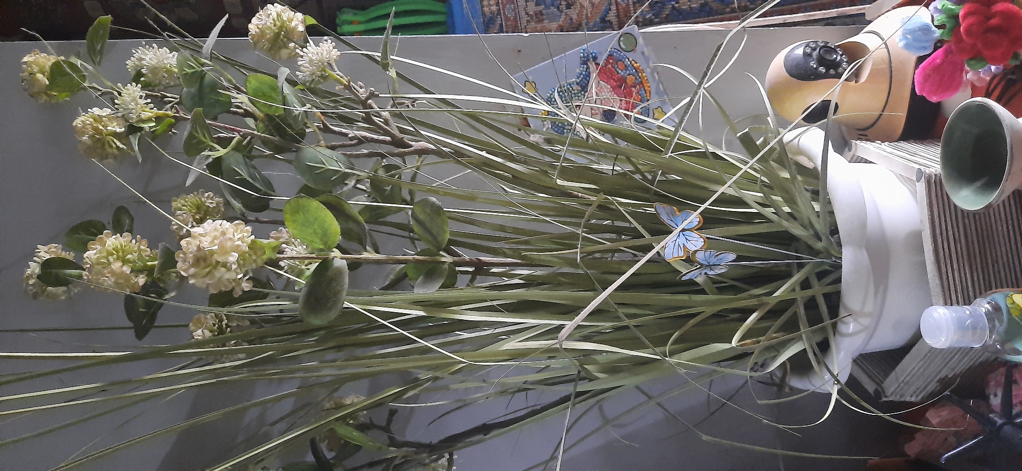 A close-up, eye-level shot shows a decorative arrangement of artificial plants and objects on a surface, possibly a shelf or table. The main focus is a large white pot filled with tall, thin, green artificial grass and flowering plants with small, off-white flower clusters. Two light blue butterflies with orange-trimmed wings are placed among the greenery. To the right of the plants, a yellow baby monitor with a black lens is visible. In the foreground, a small, clear bottle with a white cap sits on a stack of corrugated cardboard. Next to it is a small green bowl. Further to the right, brightly colored artificial flowers, primarily pink and red, are arranged. In the background, a tiled mosaic of a figure in a blue hat and red dress is mounted on a light gray wall. The lighting is soft and diffused. The overall impression is an indoor decorative arrangement with a whimsical touch. The provided crops show details of the flowers, the butterflies, the baby monitor, and the mosaic. There is no information in the image to suggest it is located in Cisauk, Indonesia.