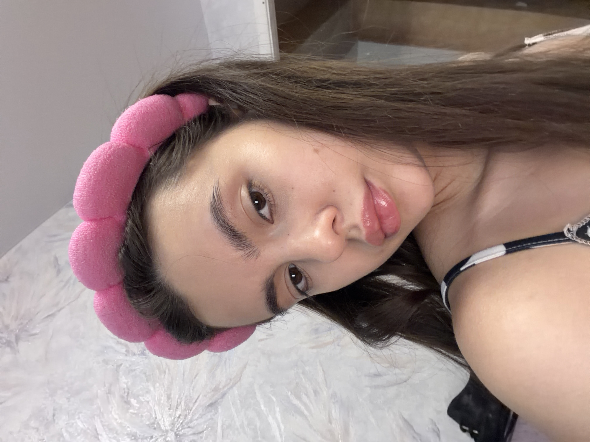 A young woman is posing for a close-up selfie. She is wearing a pink, puffy headband in her dark brown hair. She has light skin, brown eyes, and full lips. Her face is tilted slightly, and her lips are parted. A black and white striped strap is visible on her right shoulder. Part of a black object is visible in the bottom right corner. The background is a crumpled white fabric. I have no information on whether this was taken in the city of Chaikovsky, Russia.