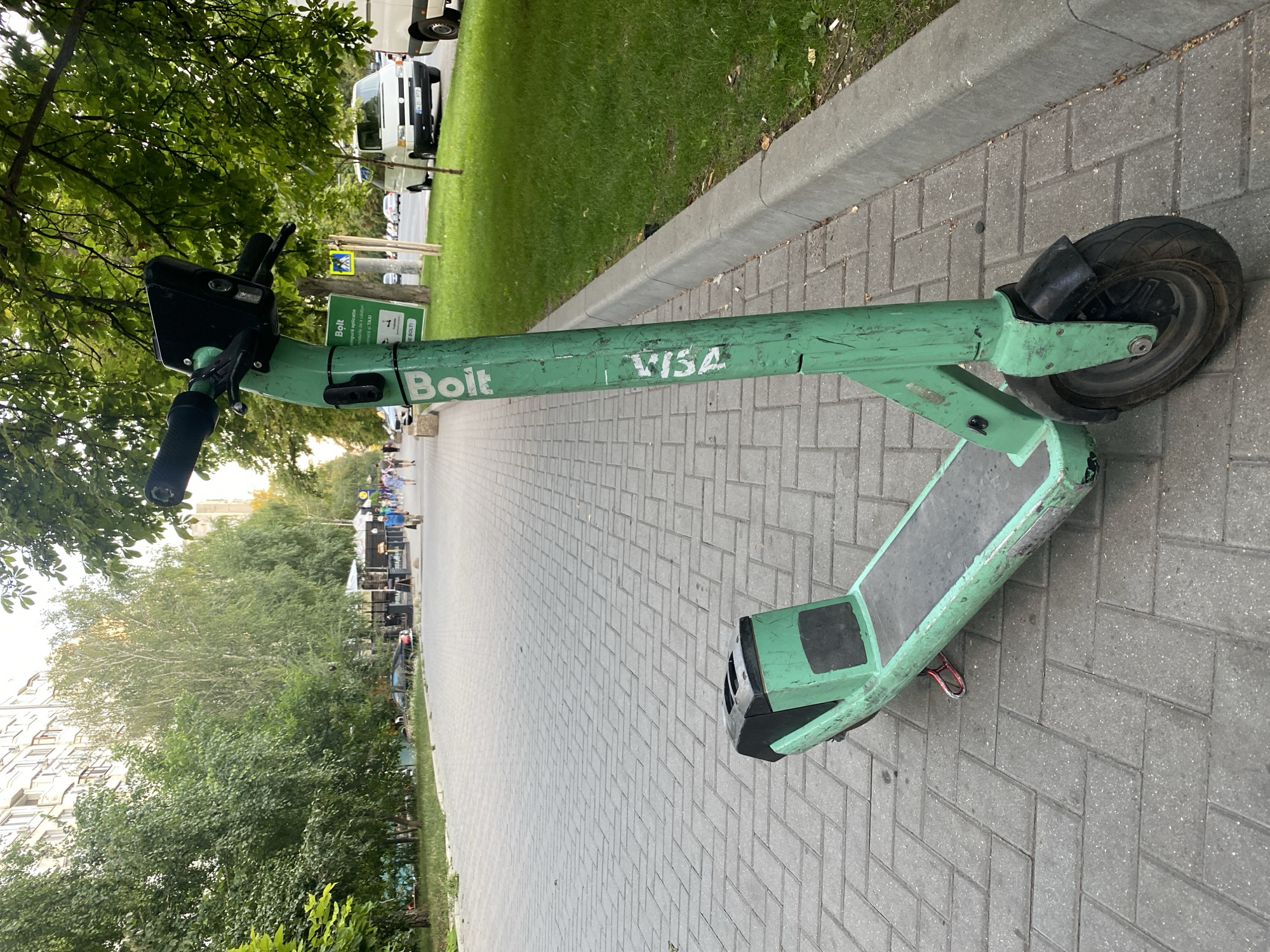 The image shows a green Bolt electric scooter parked on a paved sidewalk in Chișinău, Moldova. The scooter is standing upright, leaning slightly to its left. It has a black handlebar with a digital display and grips, and a green stem with the "Bolt" logo and "VISA" written vertically. The footboard is black and slightly worn, and there is a red kickstand visible. The scooter is positioned on a gray, rectangular paver sidewalk, with a curb and a grassy area to its right. Beyond the grass, a street is visible with several vehicles, including a white van and a black car. In the background, there are trees with green foliage and apartment buildings. Several people are walking in the distance along the sidewalk and street. The overall environment appears to be an urban street scene on a day with natural lighting.