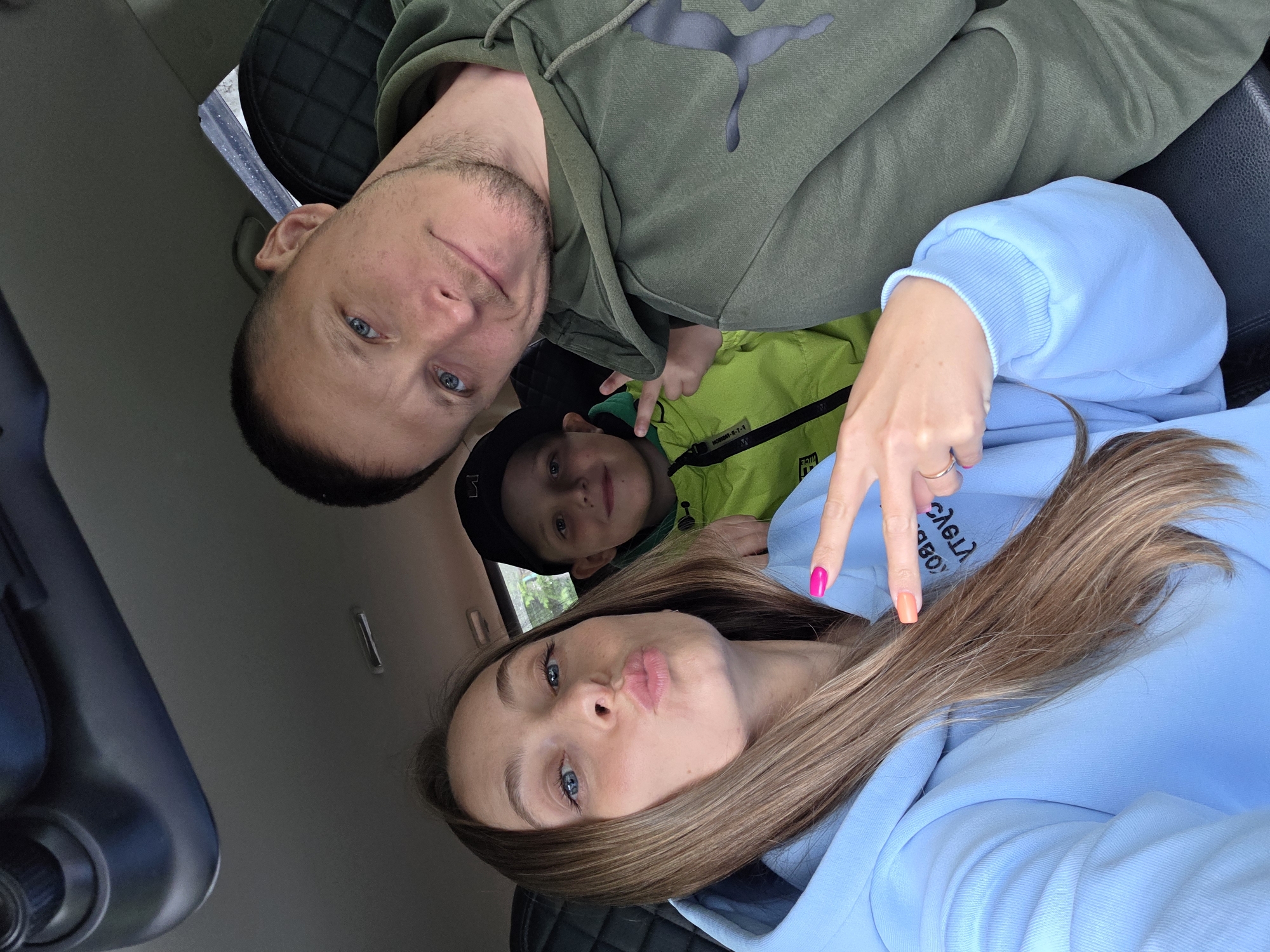 A family of three, consisting of a woman, a man, and a child, is photographed from a low-angle selfie perspective inside a vehicle in Pronkino, Russia. The woman, positioned in the left foreground, has long light-brown hair and blue eyes. She is wearing a light blue hooded sweatshirt and has pink and orange painted fingernails on her right hand, which is held up in a "V" sign. Her lips are pursed in a kissing gesture. To her right and slightly behind her is a young male child with dark hair, wearing a black baseball cap and a neon green zippered jacket. He is smiling and holds his left hand up in a "V" sign, pointing towards the man. The man, situated in the right background, has short dark hair, blue eyes, and light facial stubble. He is wearing an olive green hooded sweatshirt with a dark grey Puma logo on the left chest/shoulder area and is looking directly at the camera with a slight smile. The vehicle's interior features a grey headliner, an overhead console or rearview mirror unit at the top, and dark, possibly quilted, seat upholstery visible behind the man and child.