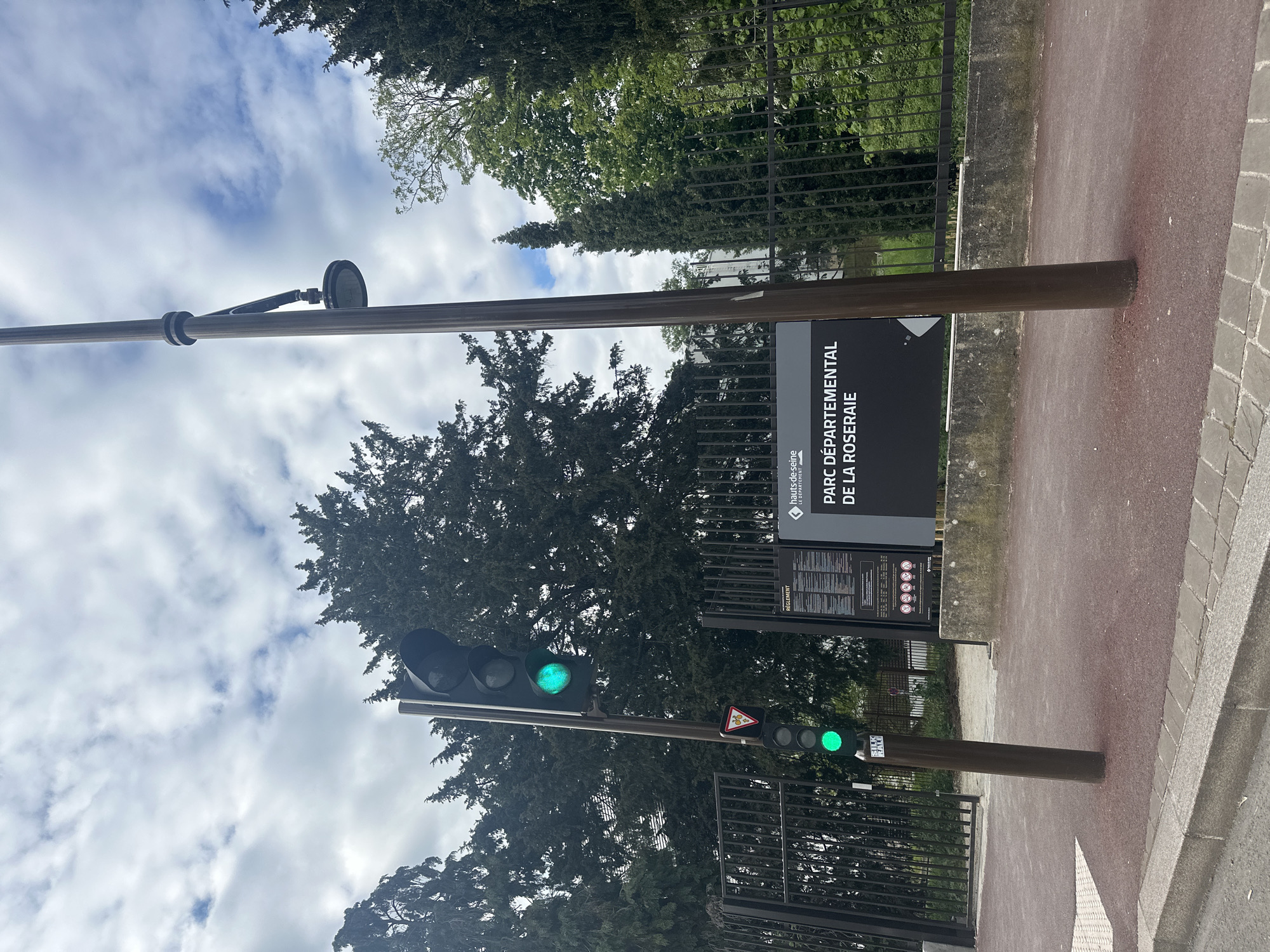 The image shows the entrance to the Parc Départemental de la Roseraie in Châtenay-Malabry, France. A sign with the park's name is visible next to a walkway. A set of traffic lights are also present, both displaying green lights. Trees are visible in the background, and the sky is cloudy.