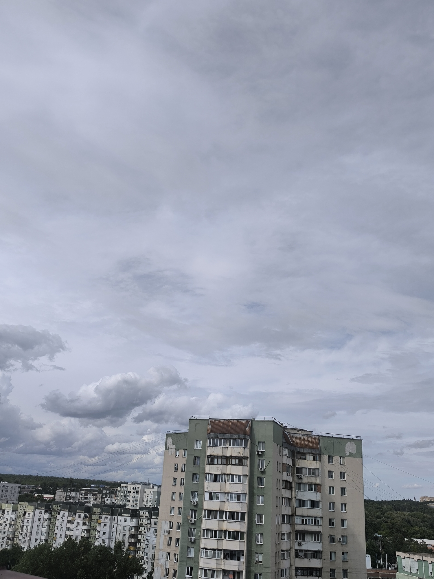 This is an image of a city skyline with a cloudy sky. The main subject is a tall, multi-story apartment building in the foreground, painted in shades of light green and grey. The building has numerous windows and balconies, and a rusted metal roof. To the left of this building, other apartment buildings are visible, extending into the distance. These buildings are also multi-story residential structures, with a variety of colors and architectural styles. A dense line of trees is visible in the lower part of the image, partially obscuring the lower floors of some buildings. In the background, a tree-covered hill slopes upwards, with more buildings visible on its crest. The sky is heavily overcast with grey clouds, suggesting a gloomy or possibly rainy day. The lighting is diffused, indicating it is likely daytime, but the lack of direct sunlight creates a muted color palette. The scene depicts an urban residential area, possibly in Chișinău, Moldova, given the location context. There is no discernible activity or event taking place; it appears to be a static view of the cityscape. No text is visible in the image.
