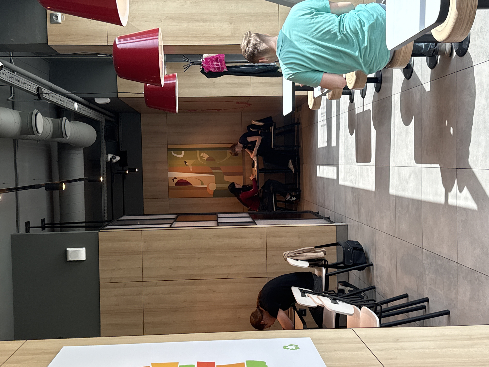The image depicts the interior of a fast-food restaurant, possibly a McDonald's, with a modern design featuring light wood paneling and a grey ceiling with exposed pipes and ventilation. In the foreground, a young man wearing a teal t-shirt is seated at a table. Further back, to the left, a person with long hair wearing a black t-shirt is seated at a table, and another person wearing a red top is seated across from them. In the background, a large abstract painting hangs on the wall. The restaurant floor is tiled, and sunlight casts strong shadows from the tables and chairs. There are also red pendant lights hanging from the ceiling and a coat rack with clothing. The overall atmosphere is casual and contemporary. This scene is located in the city of Izhevsk, Russia.
