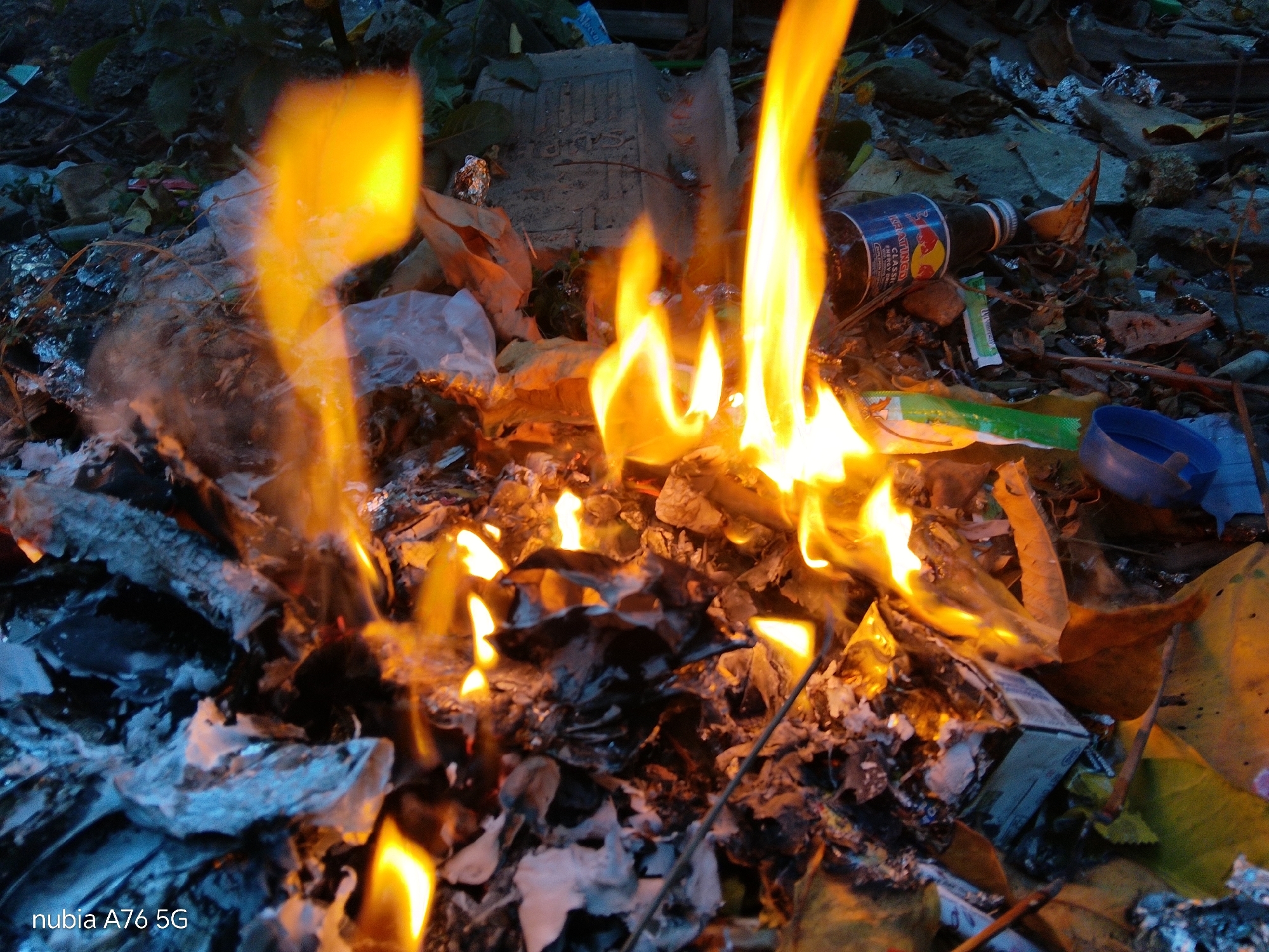 A close-up, ground-level shot depicts a small, uncontrolled fire burning amidst debris. The fire is characterized by bright orange and yellow flames, some reaching several inches in height, consuming a mixture of paper, plastic, and other assorted refuse. The surrounding area is cluttered with trash, including crumpled aluminum foil, pieces of cardboard, and what appears to be dry leaves. A partially burned bottle with a red and white label, possibly an energy drink, is visible to the right of the flames. A blue plastic lid rests near the bottle. The lighting suggests it may be dusk or a dimly lit area. The image is tagged with "nubia A76 5G", indicating the device used for capture. There is no indication of the specific location in Binuang, Indonesia, within the visual information provided.