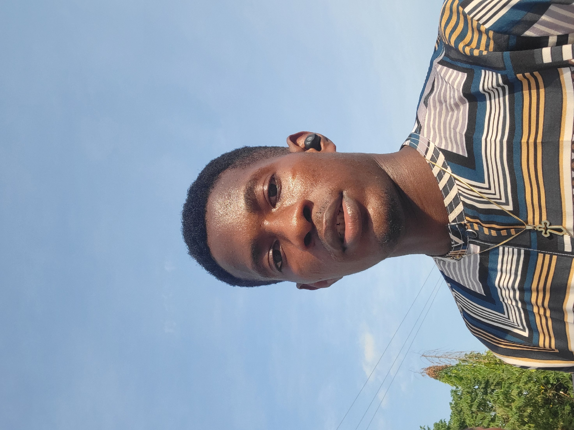 A young Black male, wearing a patterned shirt and a gold cross necklace, stands against a clear blue sky. He has short black hair and is wearing a black earbud in his right ear. The man is looking directly at the camera. Trees and utility lines are visible in the background, suggesting an outdoor setting in Egbagu, Nigeria. The lighting indicates it is daytime.